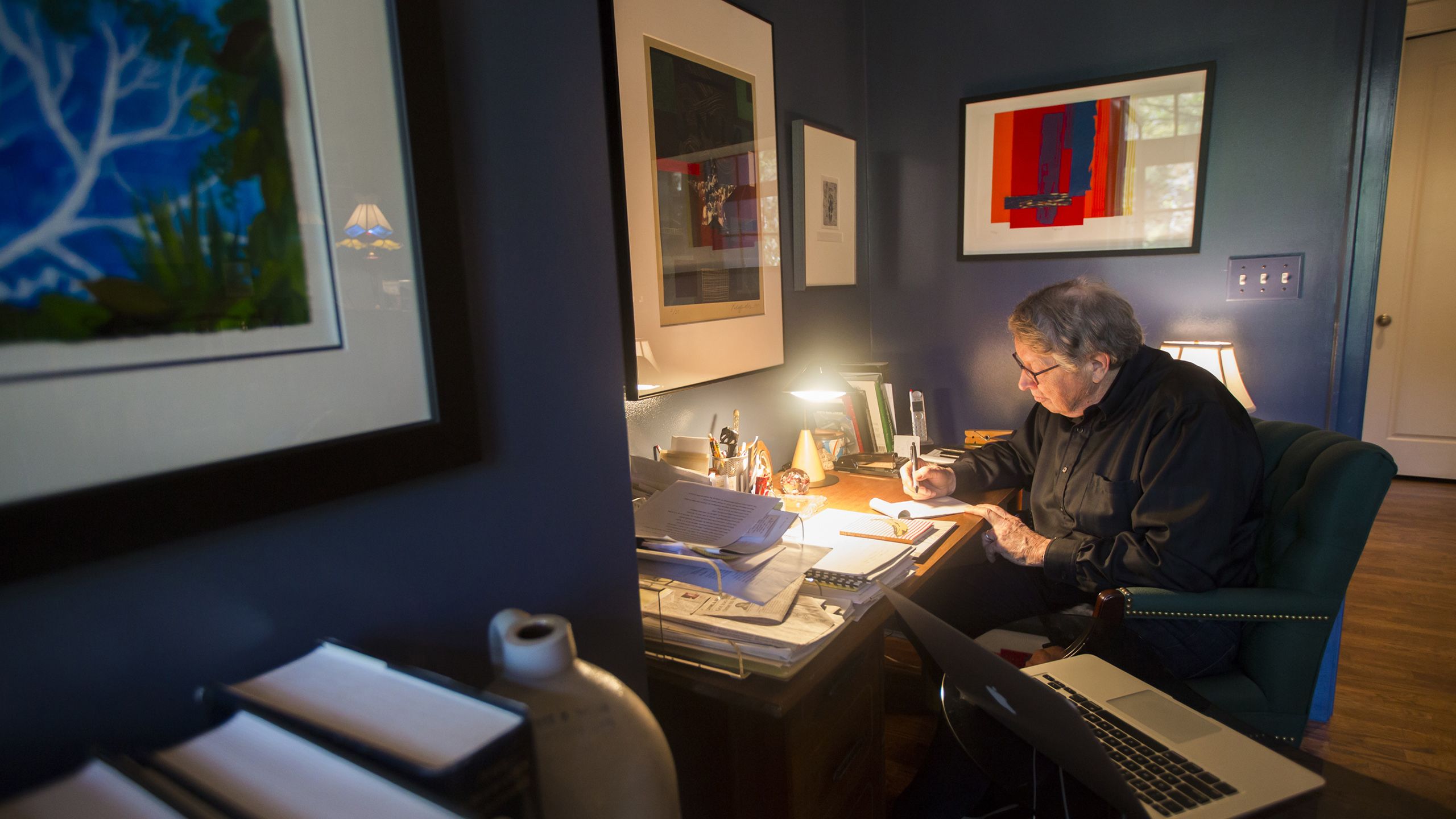 Burkett sitting at his desk writing a note, surrounded by papers with artwork on the walls