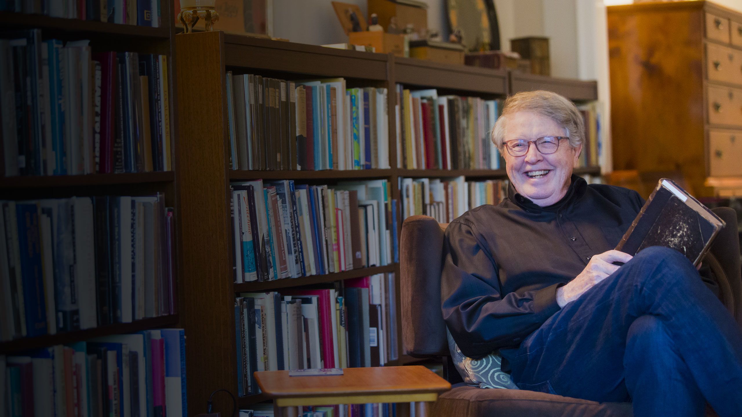 Burkett smiles while sitting in a chair in his home library holding a book