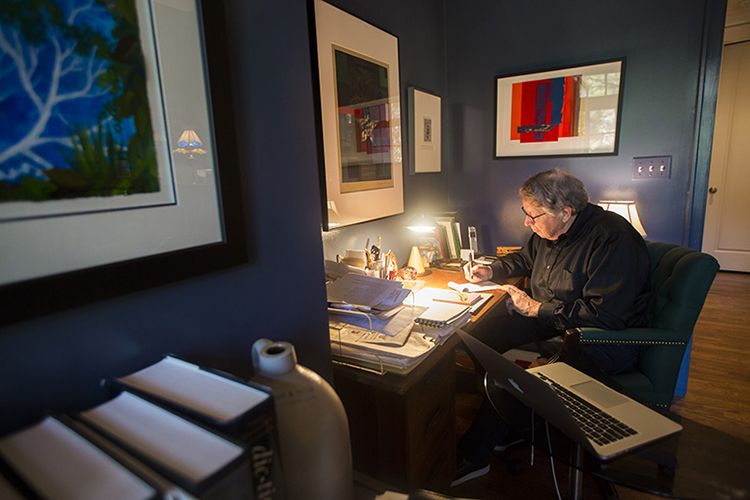 Burkett writing a note at his desk. Framed art hangs on the walls surrounding his desk.