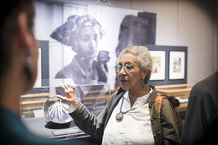 Camille Billops standing in the gallery with her hand resting on the top of a display case containing her ceramics