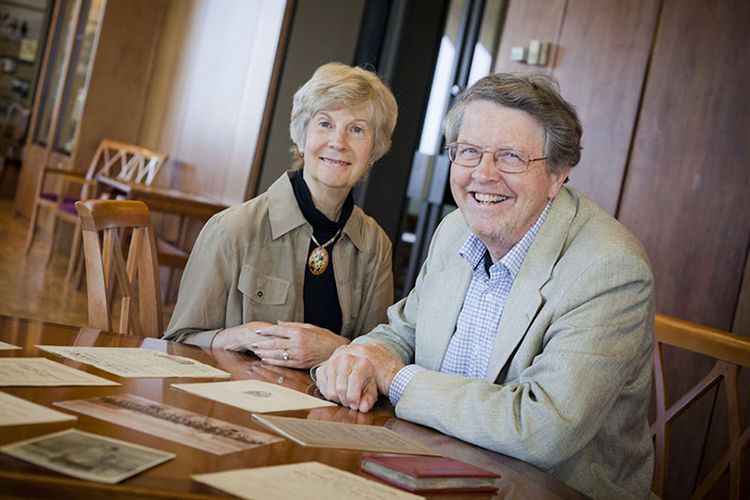 Nancy Burkett and Randall Burkett smiling while sitting next to each other at a wooden table in Rose Library. Several papers and books are resting on the table in front of them.