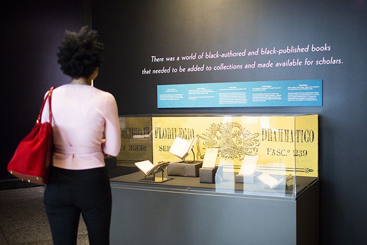 A woman standing facing a display case in front of a wall with a vinyl quote that reads “There was a world of Black-authored and Black-published books that needed to be added to the collection and made available for scholars.”