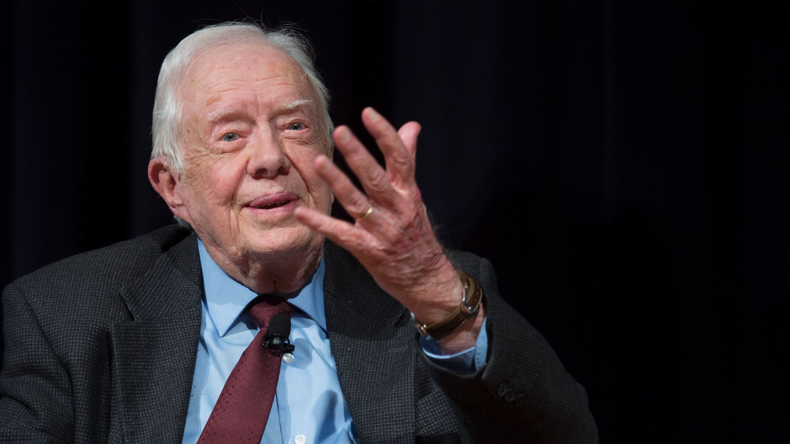 President Carter speaks with his hand raised, wering a dark suit, light blue shirt and burgundy tie.