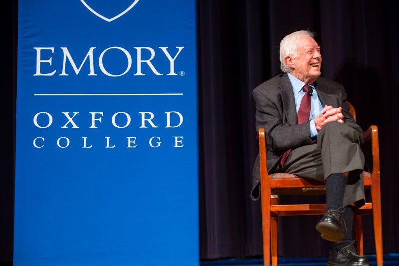 President Carter sits on a chair on stage by a blue banner that reads "Emory Oxford College."