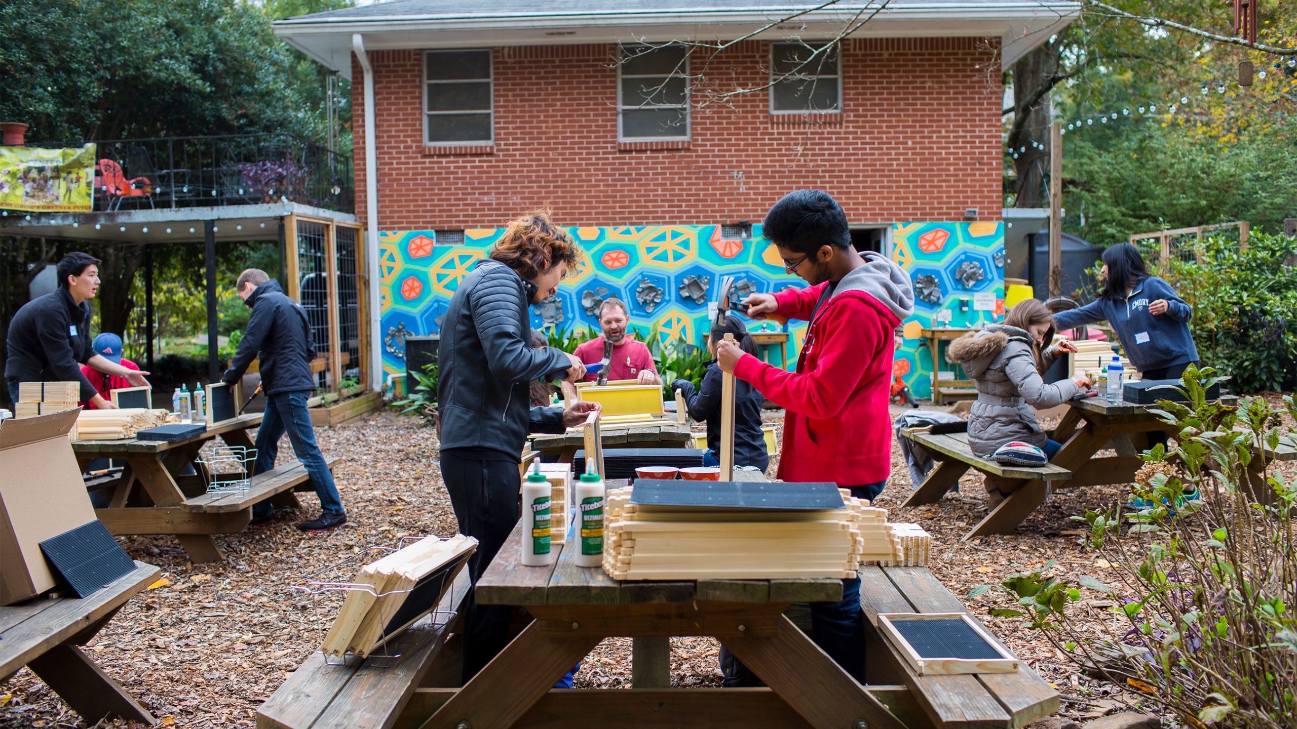 Students work on a building project outside on Emory Cares Day. They are shown hammering wood at a picnic table in front of a bright mural.