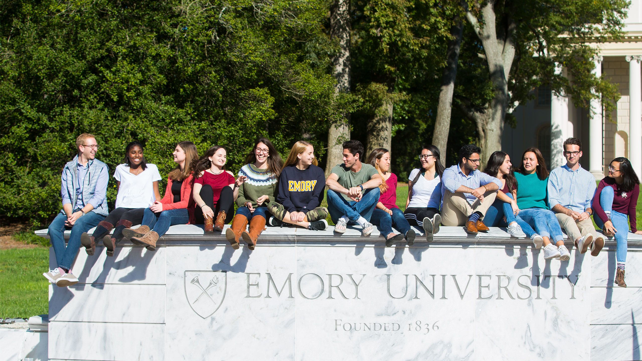 A diverse group of students sits on top of the white marbel wall that markes the entrance to Emory University. The Emory shield and name are carved in the wall.