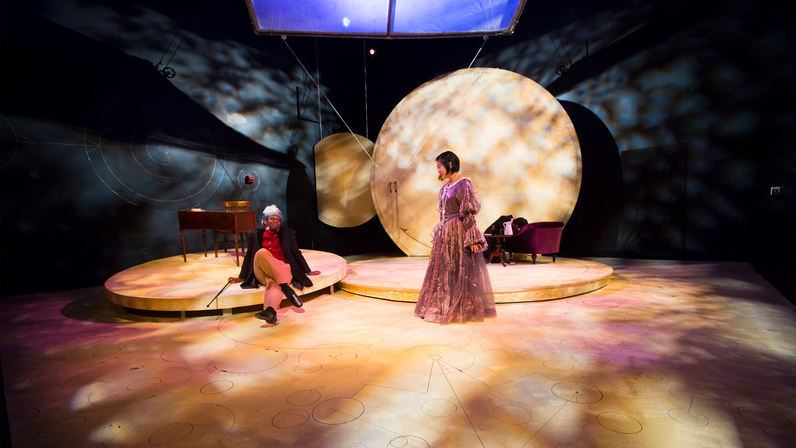 Two actors in 1800s period costumes stand on a stage at Oxford College.