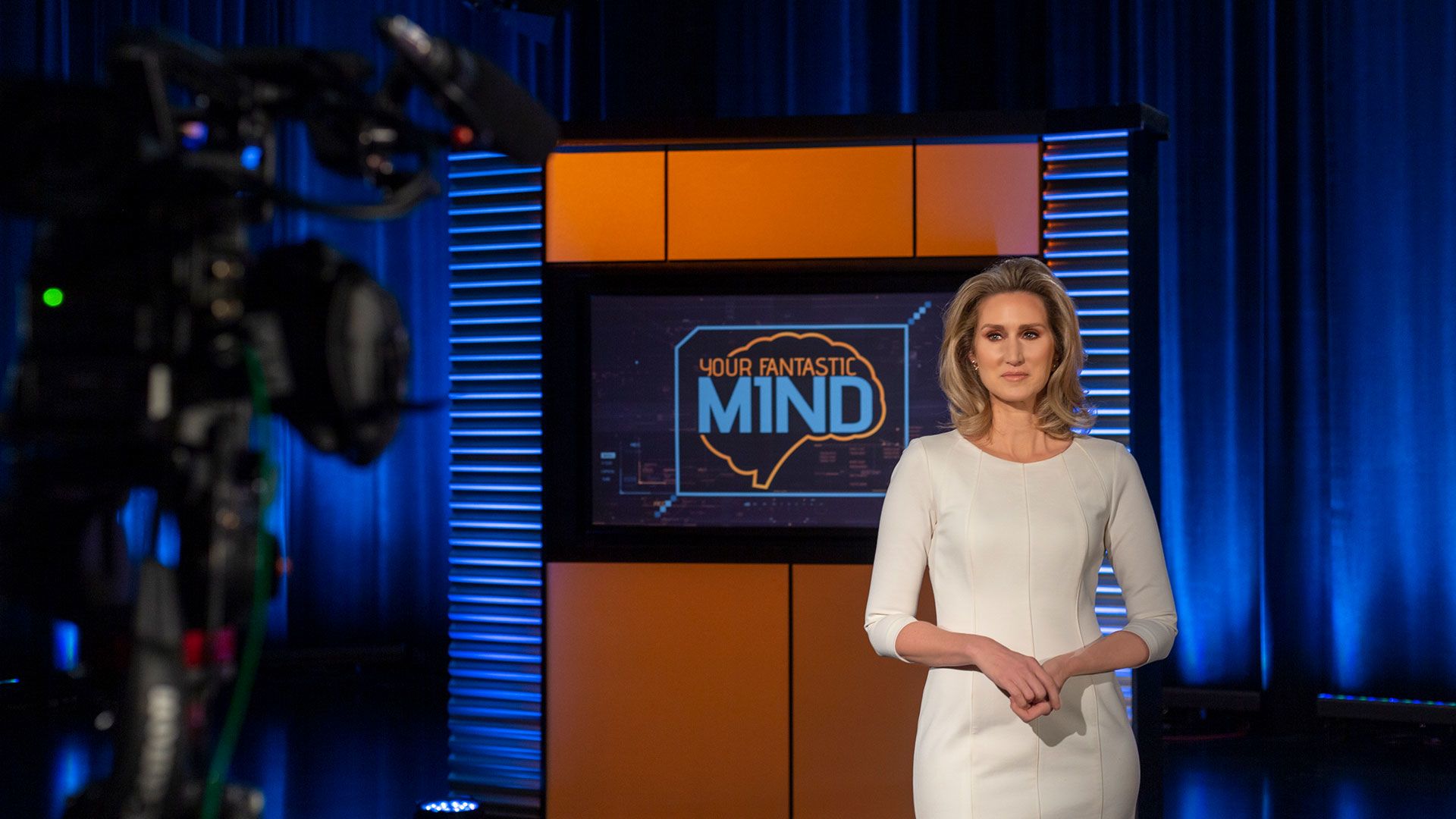 Your Fantastic Mind host Jaye Watson stands in front of a logo of the show on the studio set. 