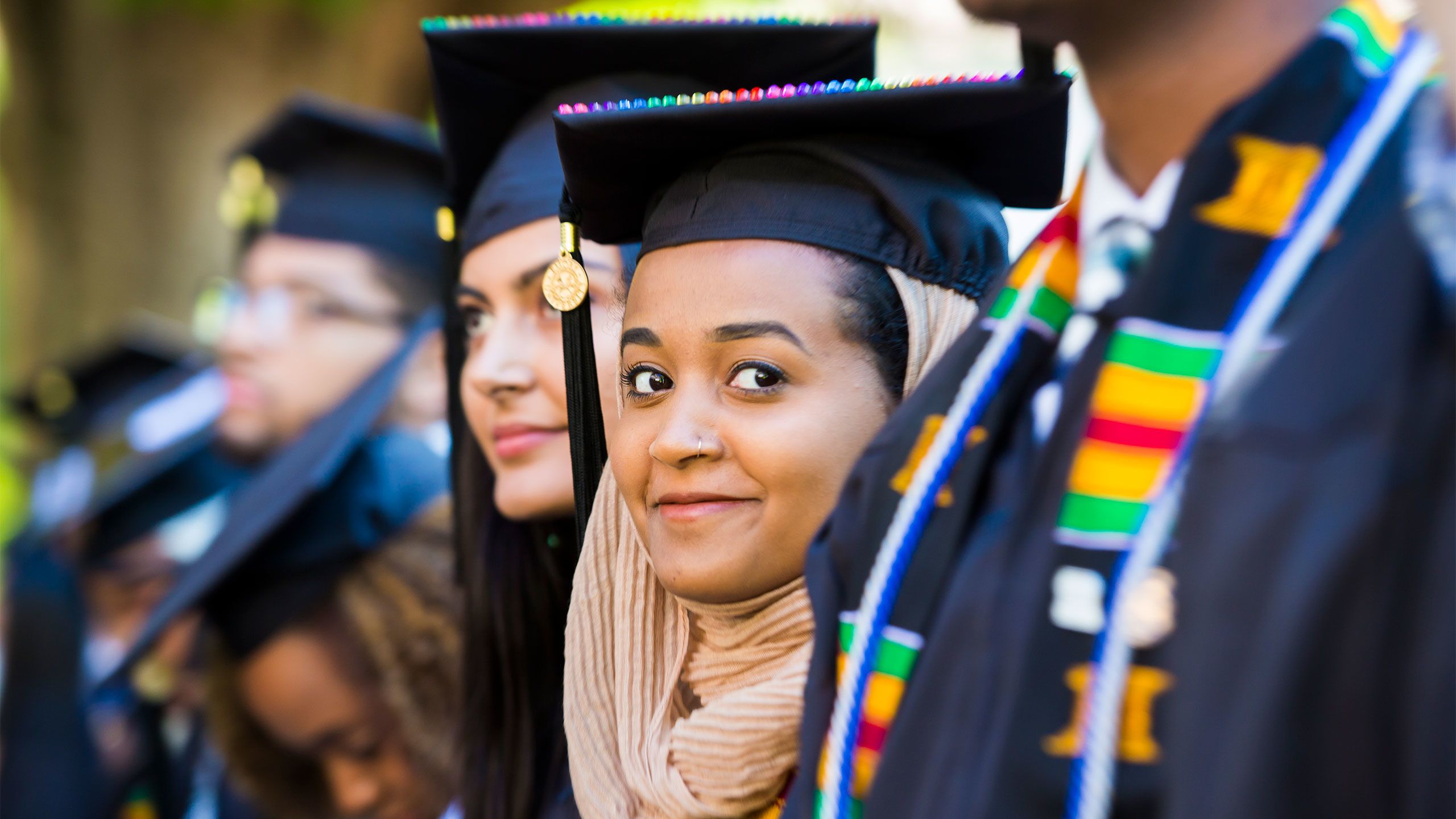 A young woman wearing a head scarf with her graduation cap smiles at the camera