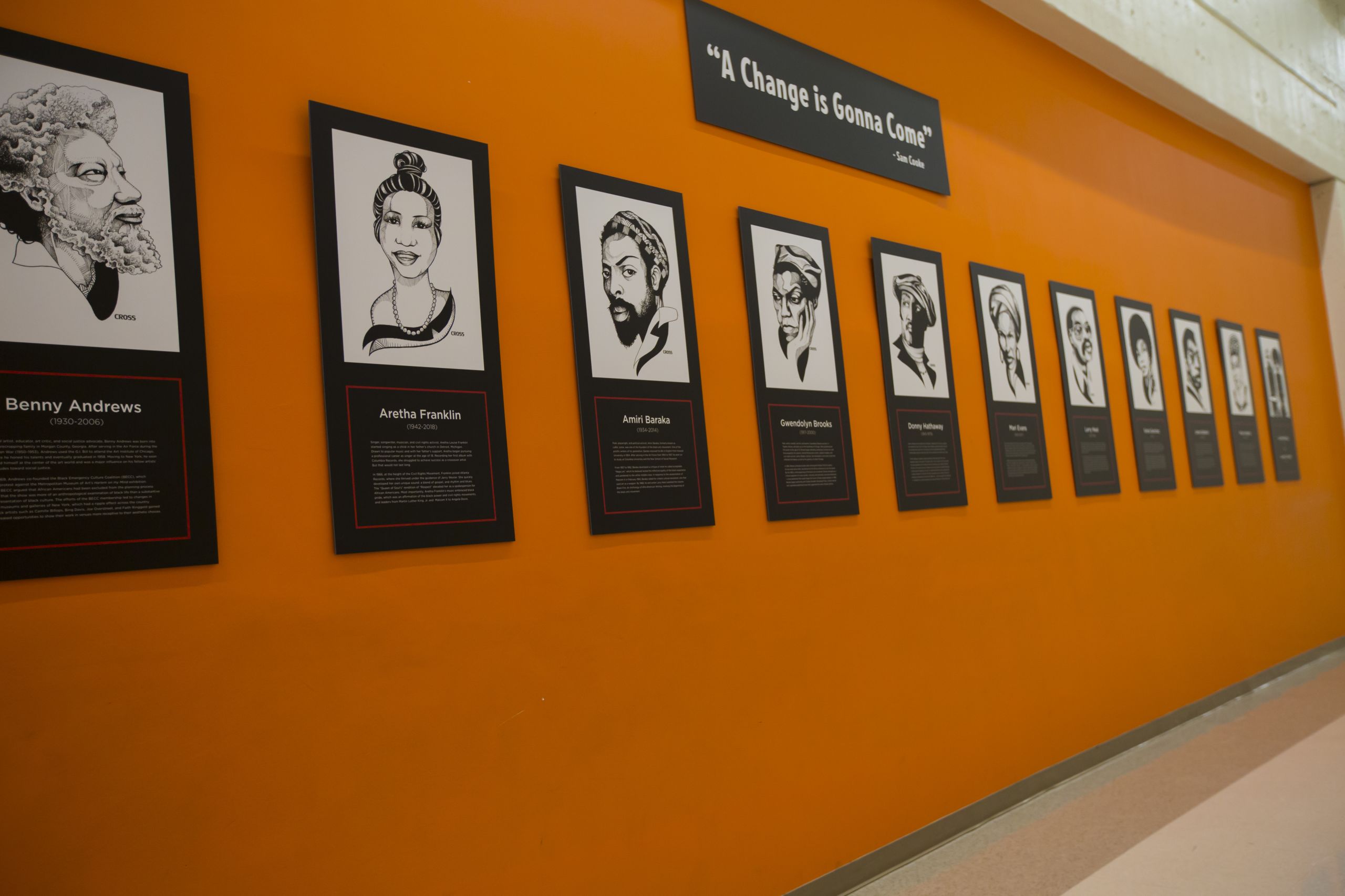 A hallway painted orange has the quote "A Change is Gonna Come" above posters featuring black and white drawings of African American arts leaders.