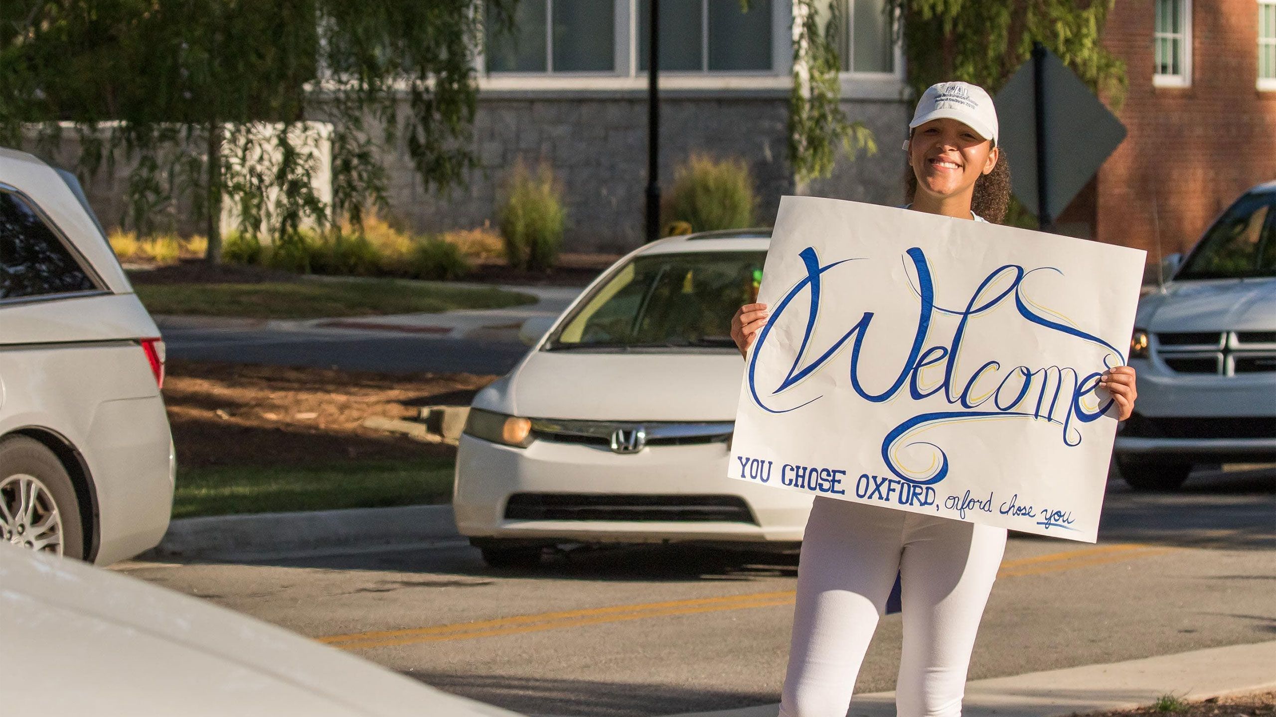 A student holds a handwritten "Welcome" sign as cars stream in at Oxford College
