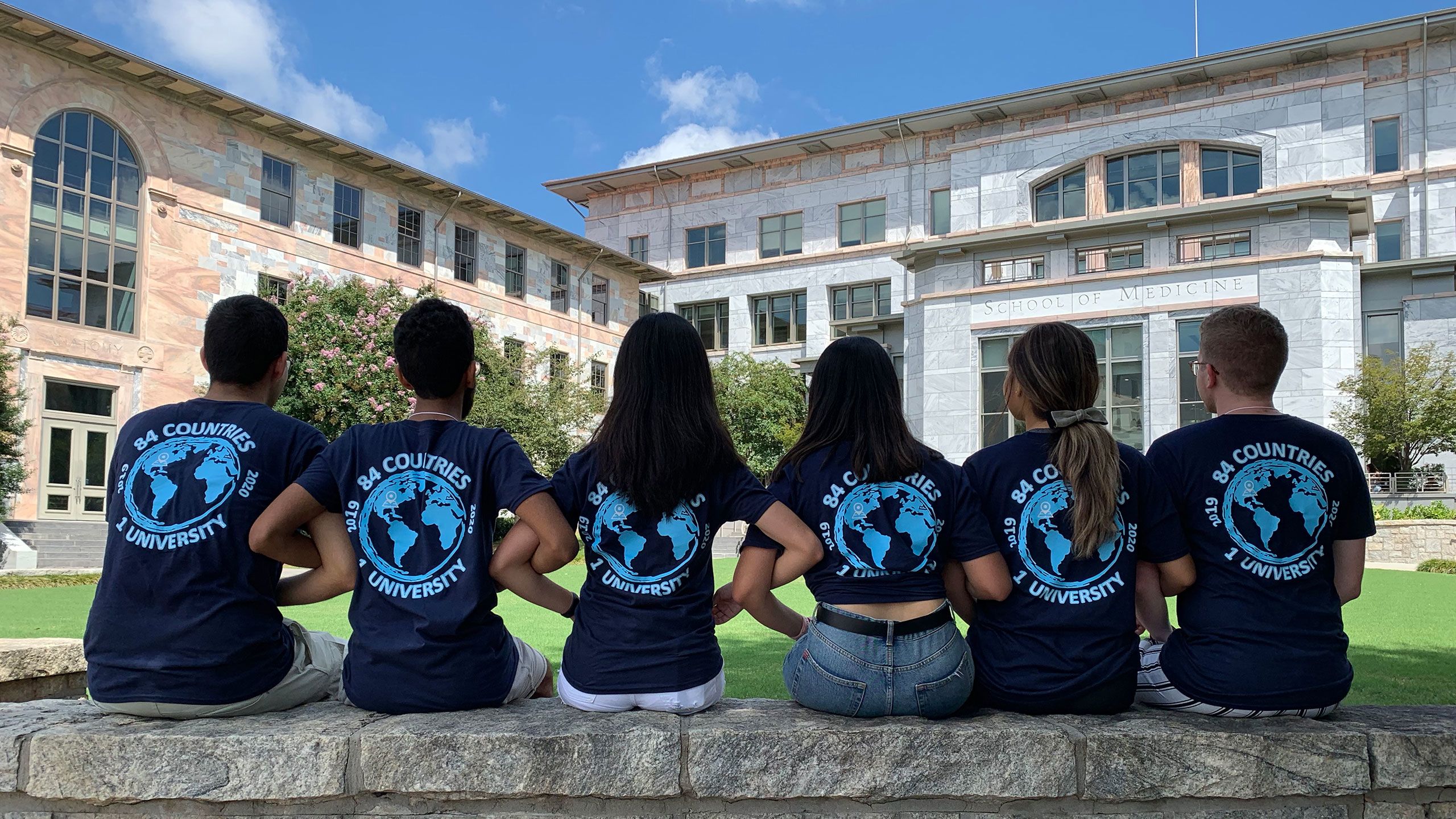 Students sit on a wall with their arms linked. The backs of their t-shirts say "84 Counties - 1 University"