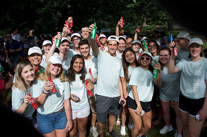 A large group of students in white shirts hold Coca-Cola aluminum bottles in the air.