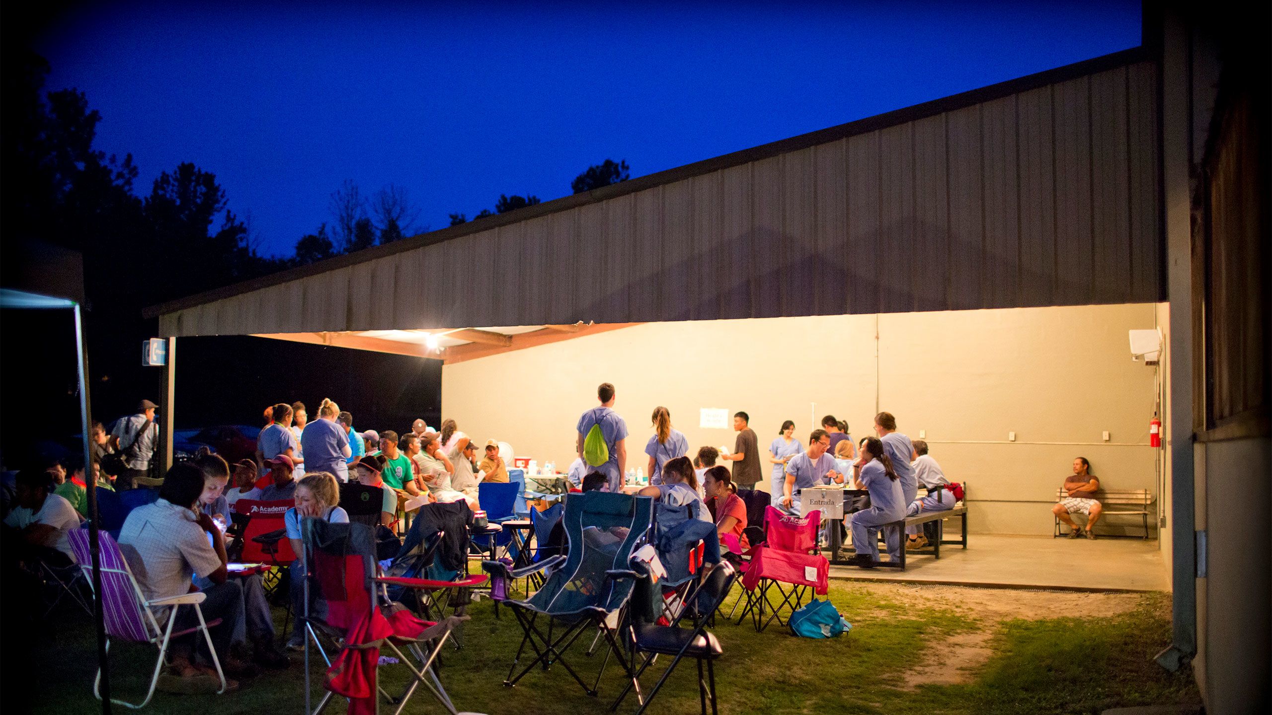 farm workers and health students sit in camp chairs and at picnic tables at a night health clinic