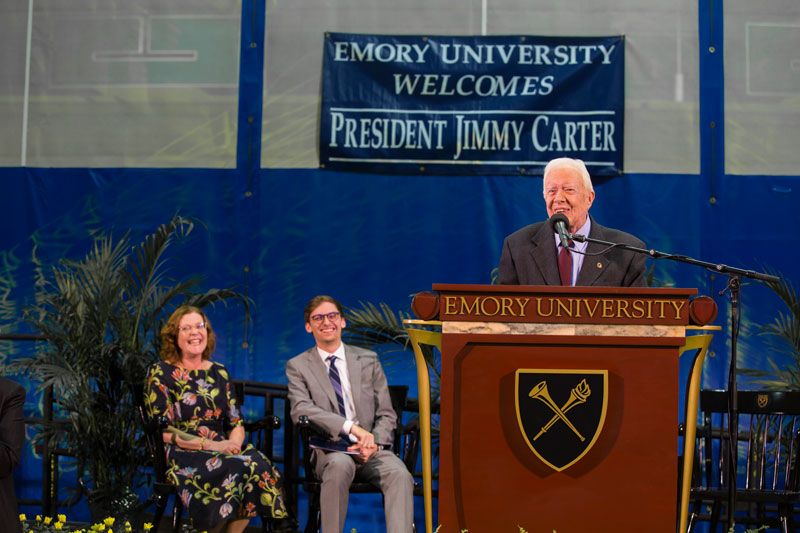 Emory President Claire E. Sterk and Student Government Association President Ben Palmer sit in chairs on the stage where U.S. President Jimmy Carter stands at the podium.
