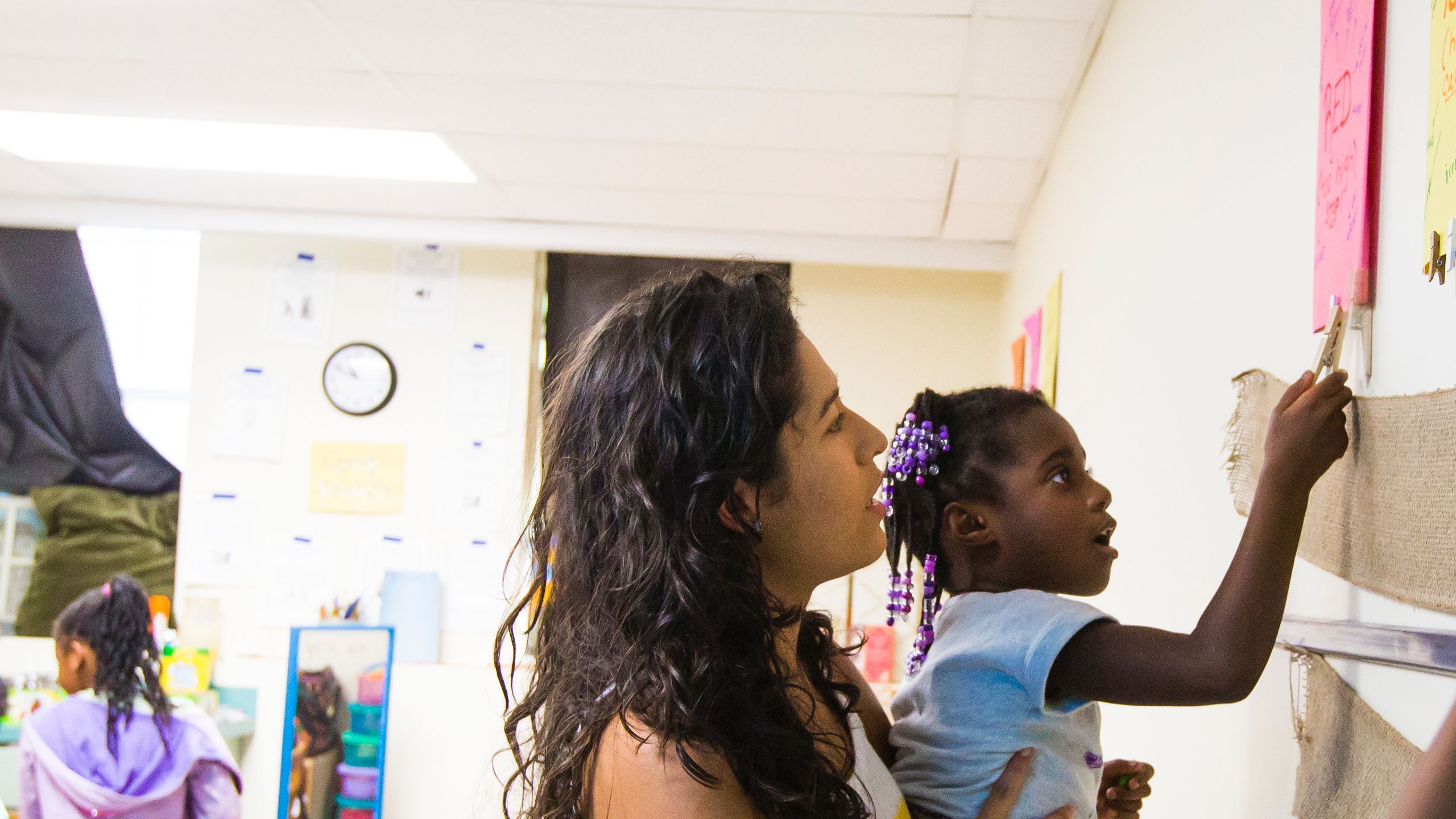 College student holds up a child to reach things on the classroom wall