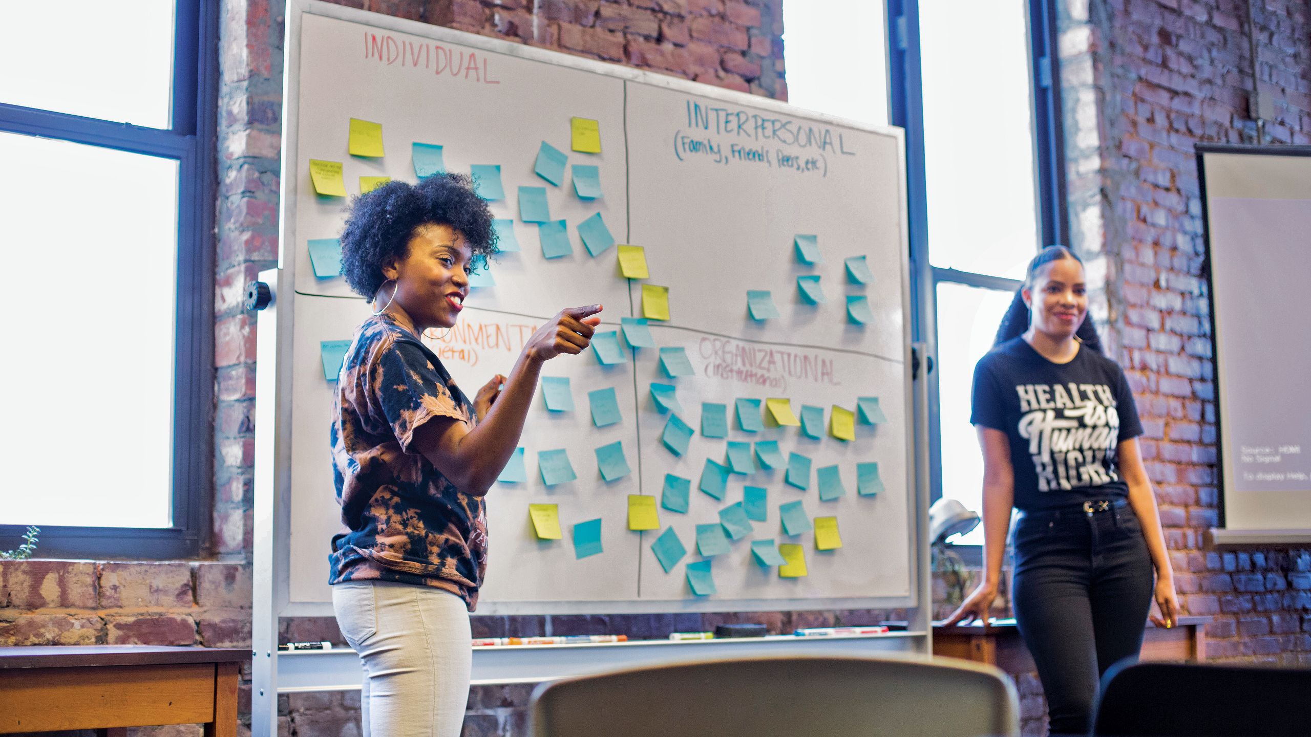 Showing Khadijah Ameen and Mercilla Ryan-Harris stand in front of a white board covered with blue and  yellow Post-It notes that contain attendees' comments.