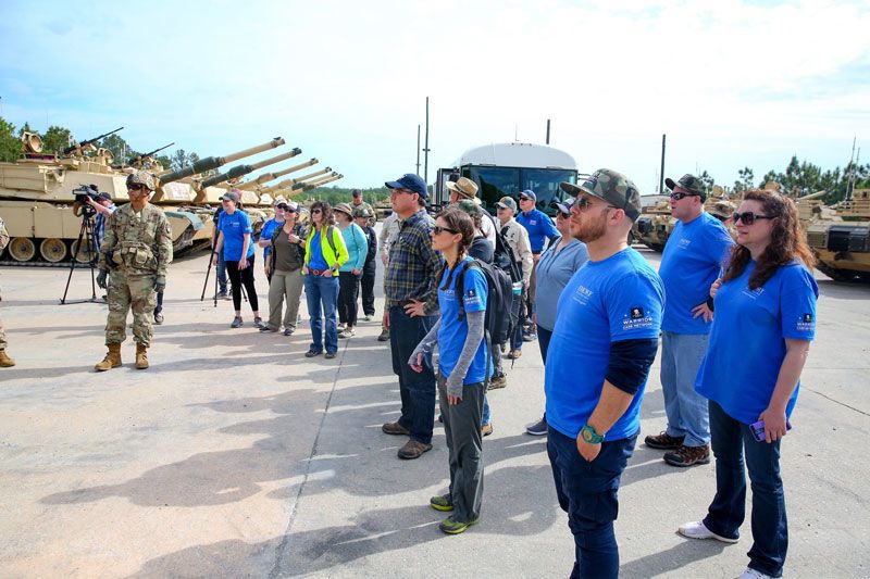 Emory Healthcare leaders wearing blue shirts stand with soldiers near a line of tanks at Fort Benning.
