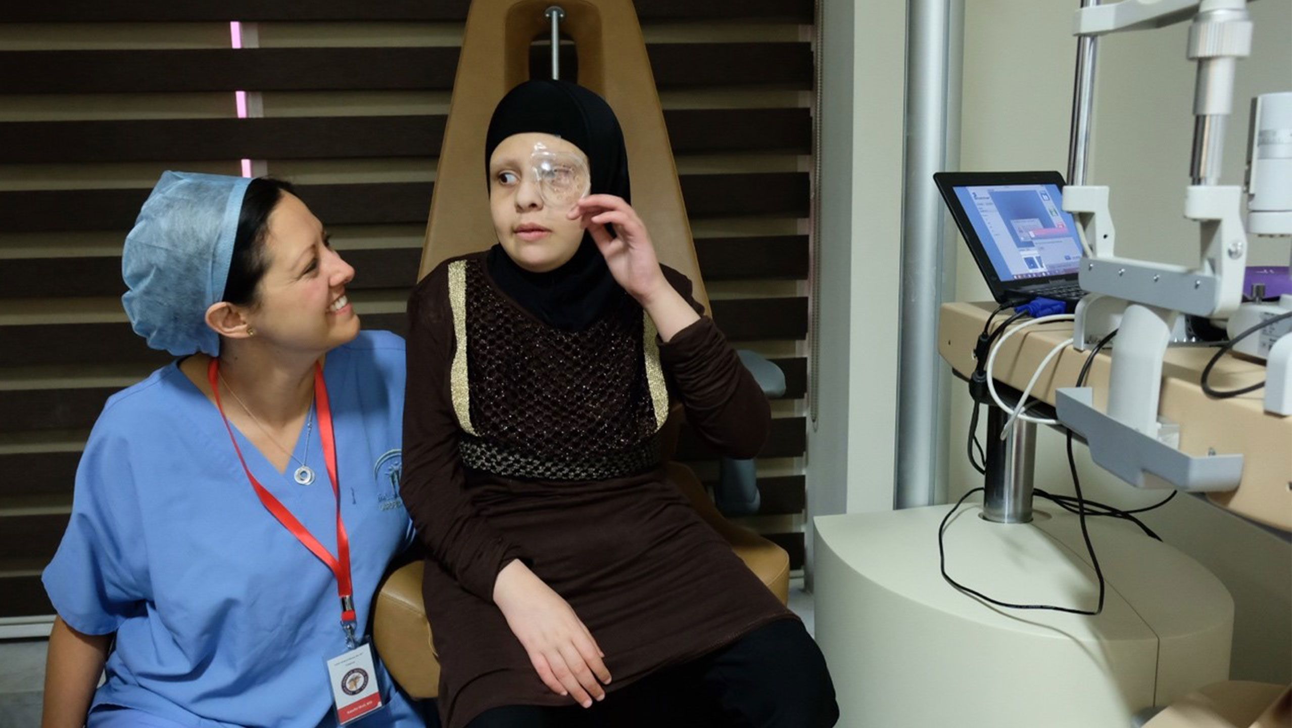An Emory physician in blue scrubs talks with a young Syrian girl wearing an eye patch