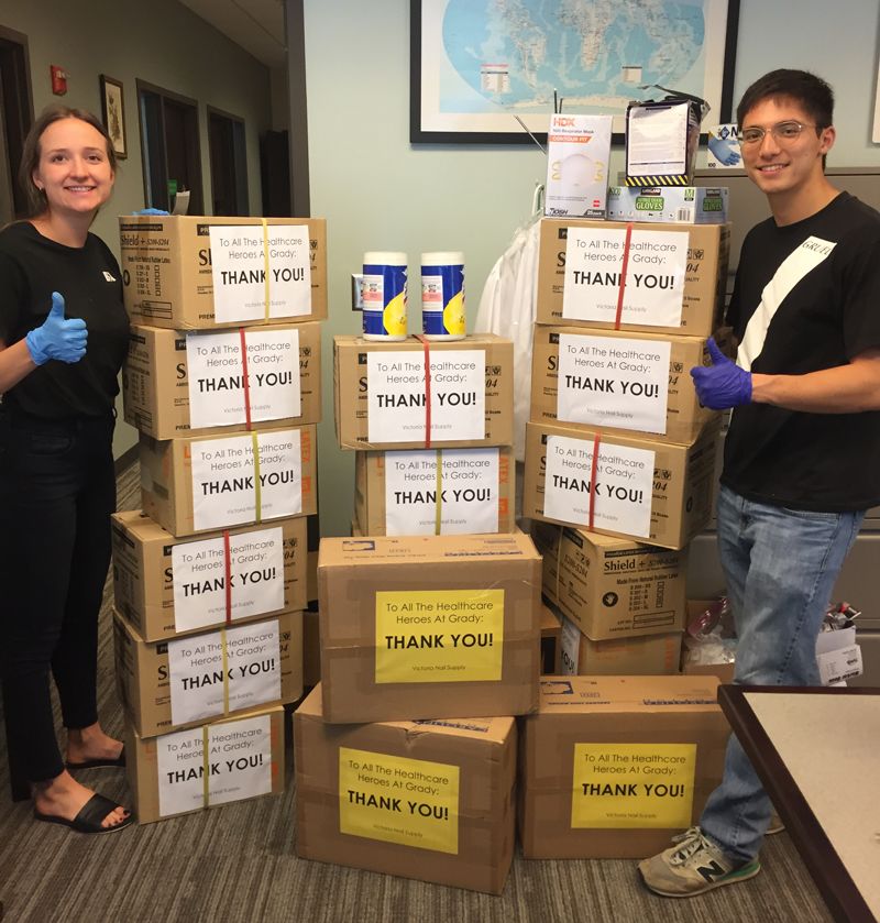 Two students wearing gloves and standing apart give a "thumbs up" while posing with boxes of donated medical supplies that have signs thanking health care workers for their service.
