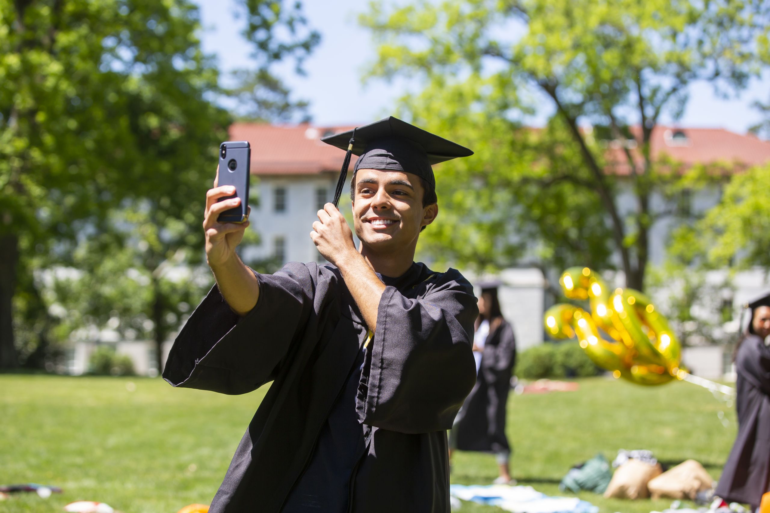 A student watches Commencement on his cell phone as he moves the tassel on his cap from right to left