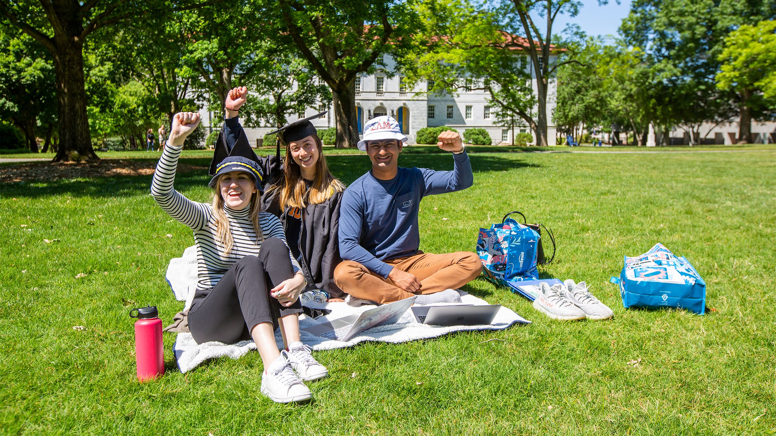 A student in cap and gown sits on a blanket on the quad watching Commencement on a laptop