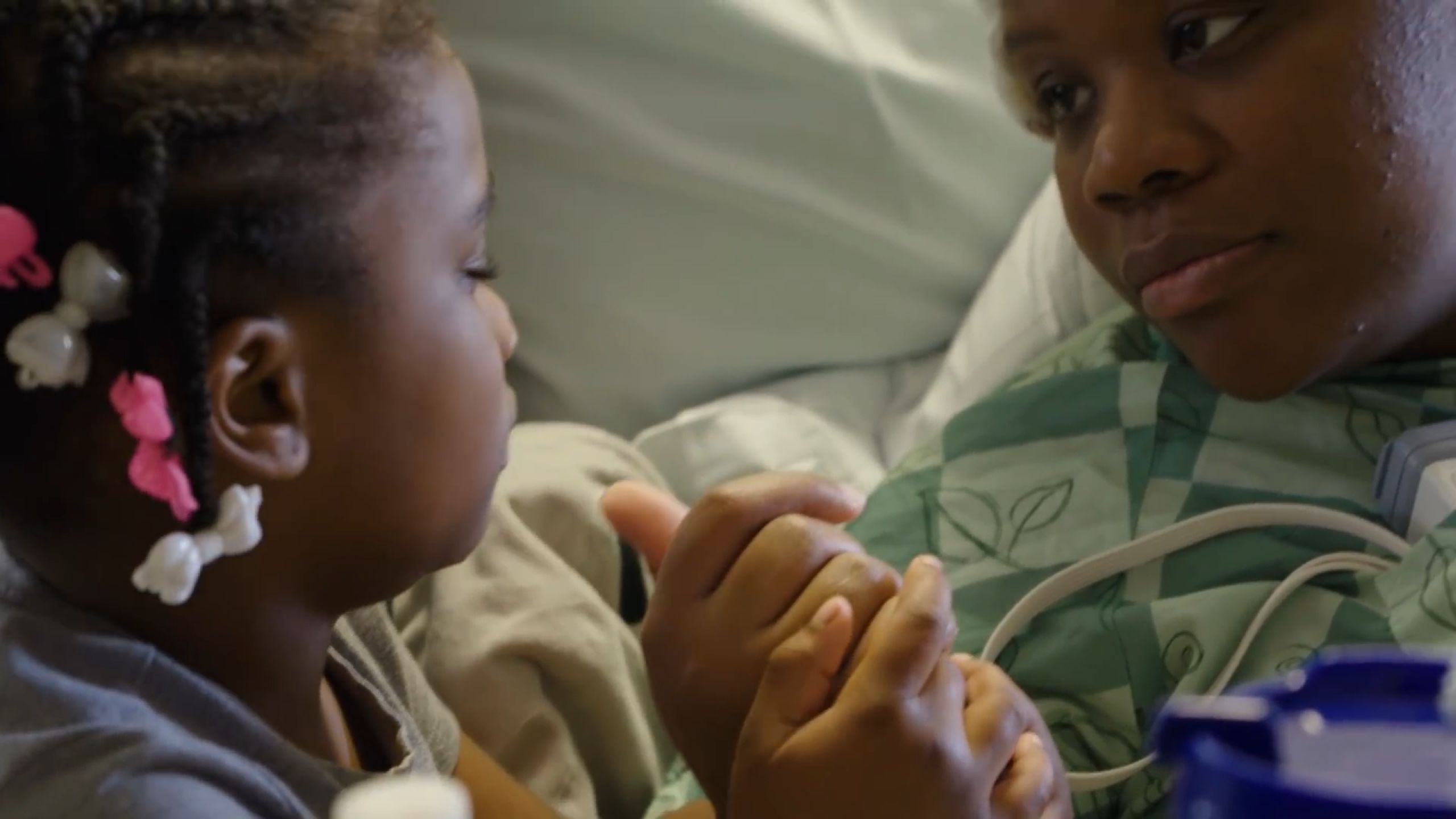 An epilepsy patient in a hospital bed and gown holds her young daughter's hand.