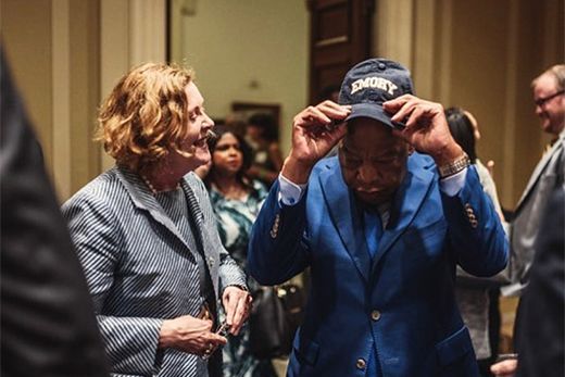 John Lewis places a blue Emory baseball cap on his head as Emory President Claire Sterk looks on smiling