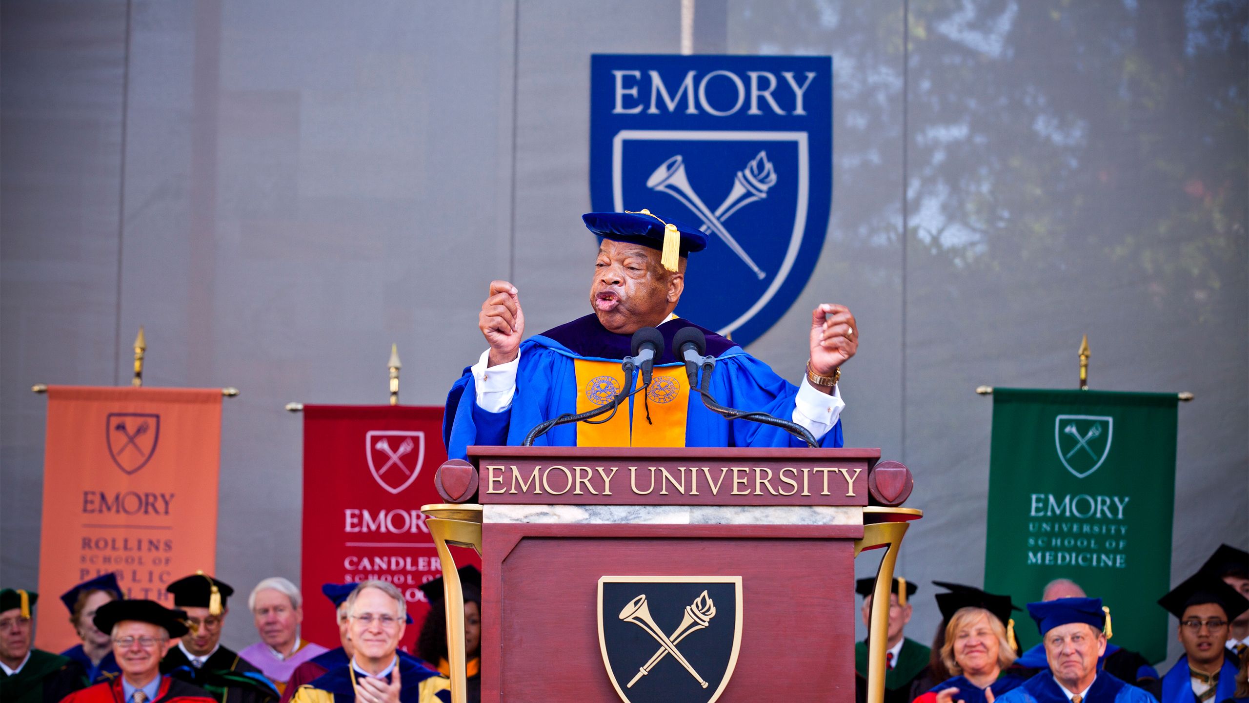 Wearing academic robes, U.S. Rep. John Lewis speaks at an Emory podium with the Emory shield in the background
