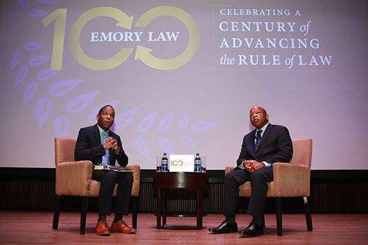 Fred Smith Jr. and John Lewis sit in chairs on a stage. A backdrop notes 100 years of Emory Law with the words "Celebrating a Century of Advancing the Rule of Law"