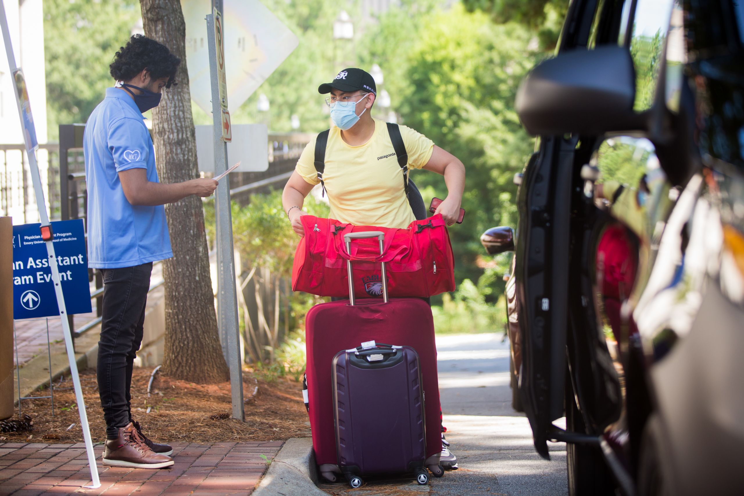 A student wearing a mask is greeted as he carries a suitcase and duffle bag
