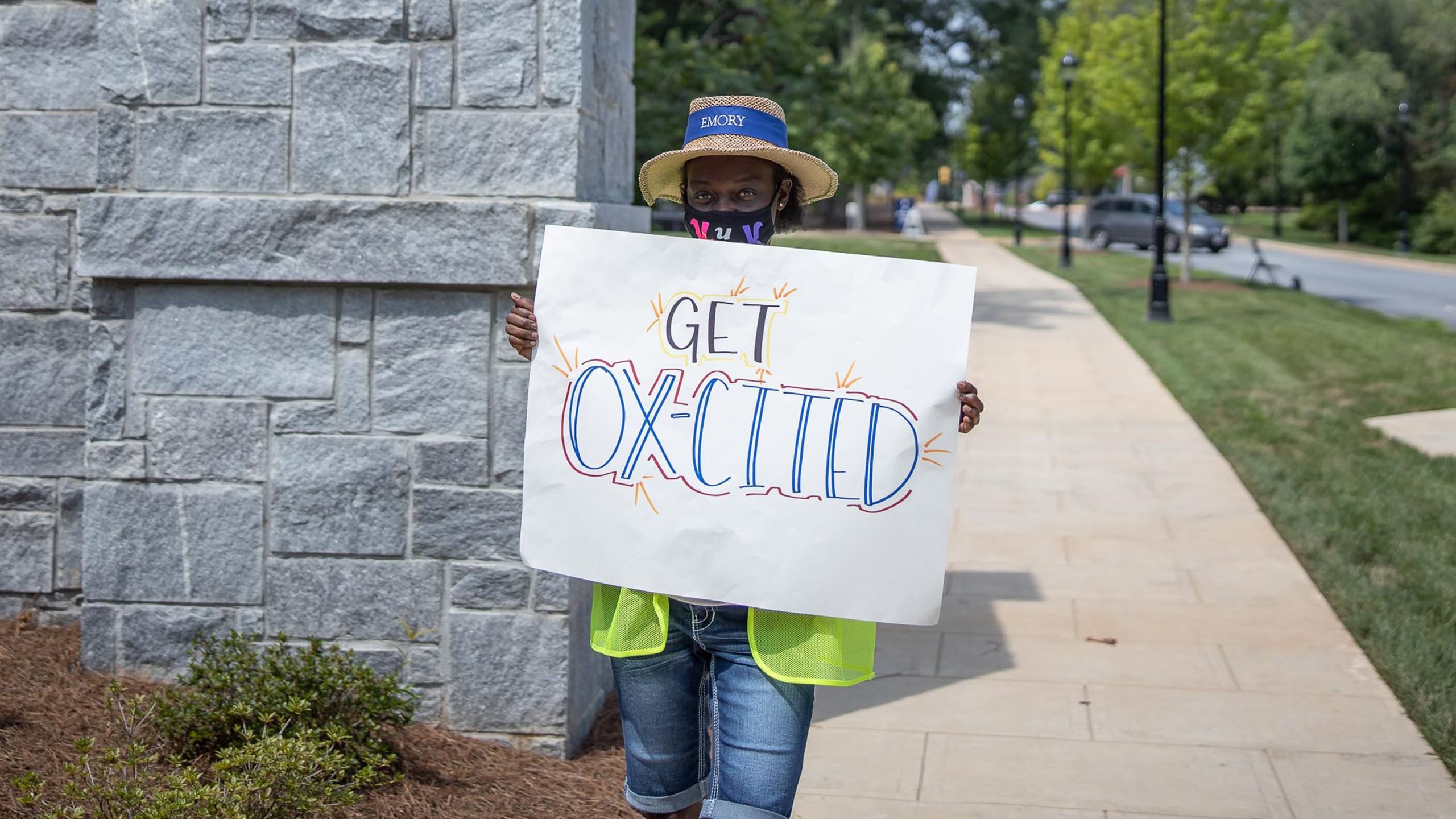 A woman in a face covering and Emory hat holds a welcome sign at the entrance to Oxford College