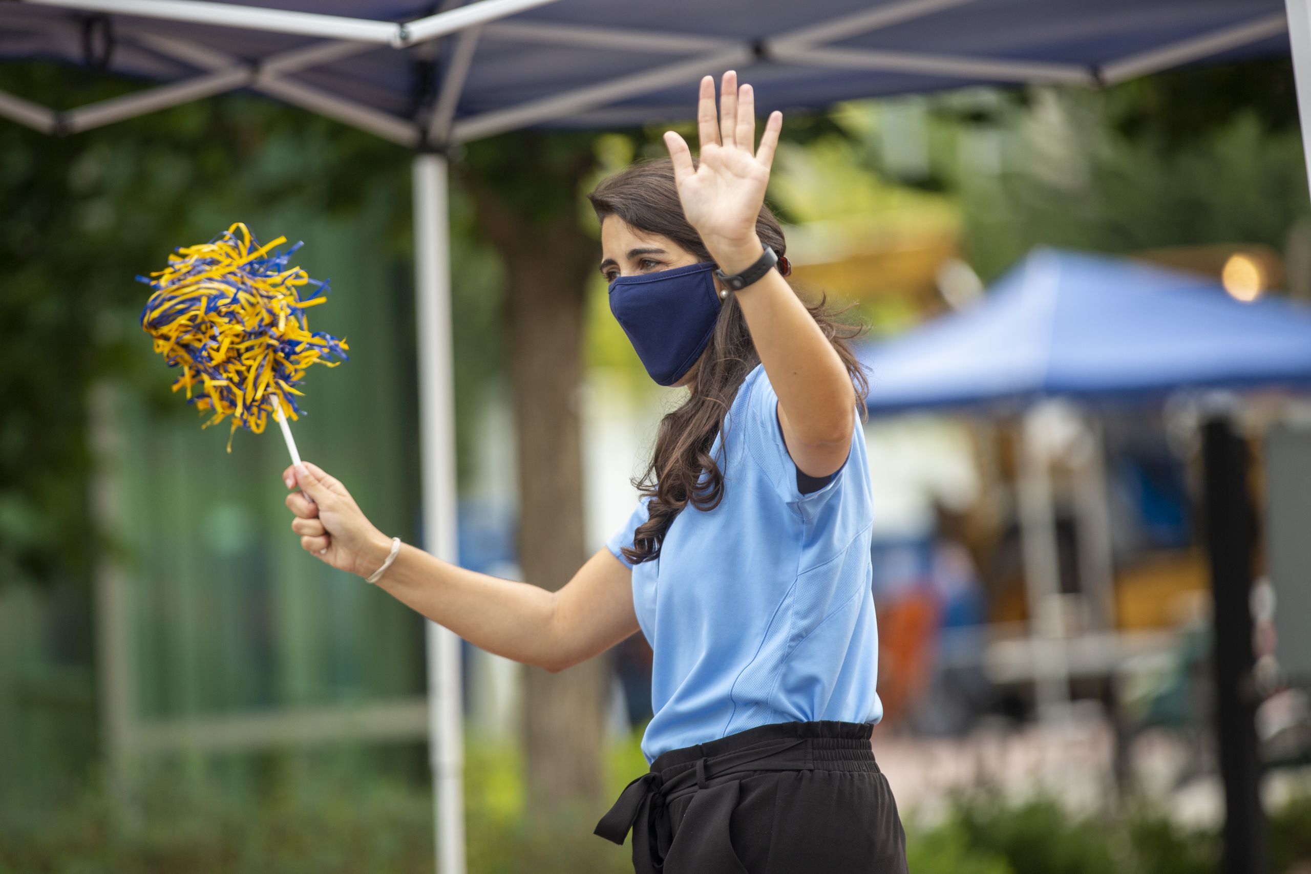 A woman in a face mask and blue shirt waves a blue and gold pom-pom to welcome students