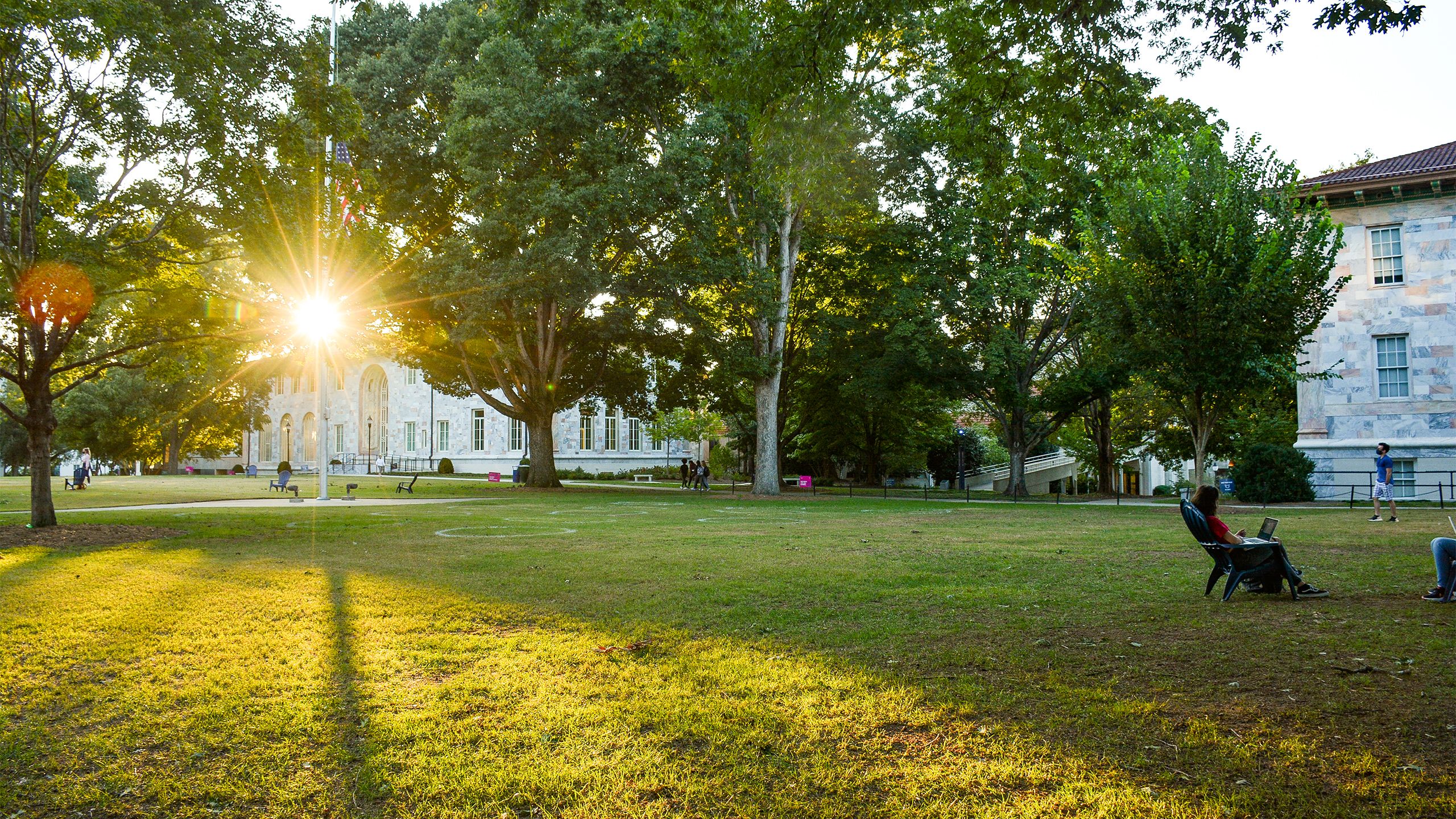 A view of the Emory Quad at sunset shows students scattered in chairs and the sun shining through the trees