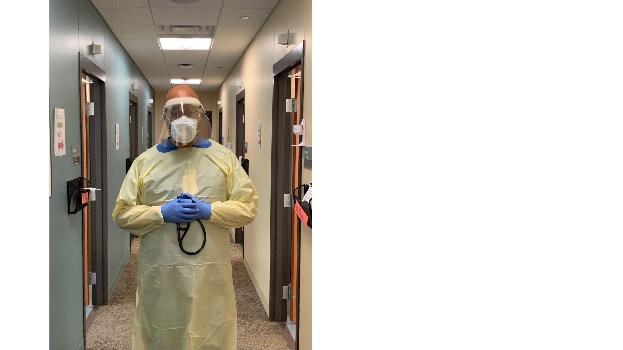 A doctor stands in the hallway of the clinic where he works in full personal protective equipment (PPE) including gown, gloves, mask, and face guard. 