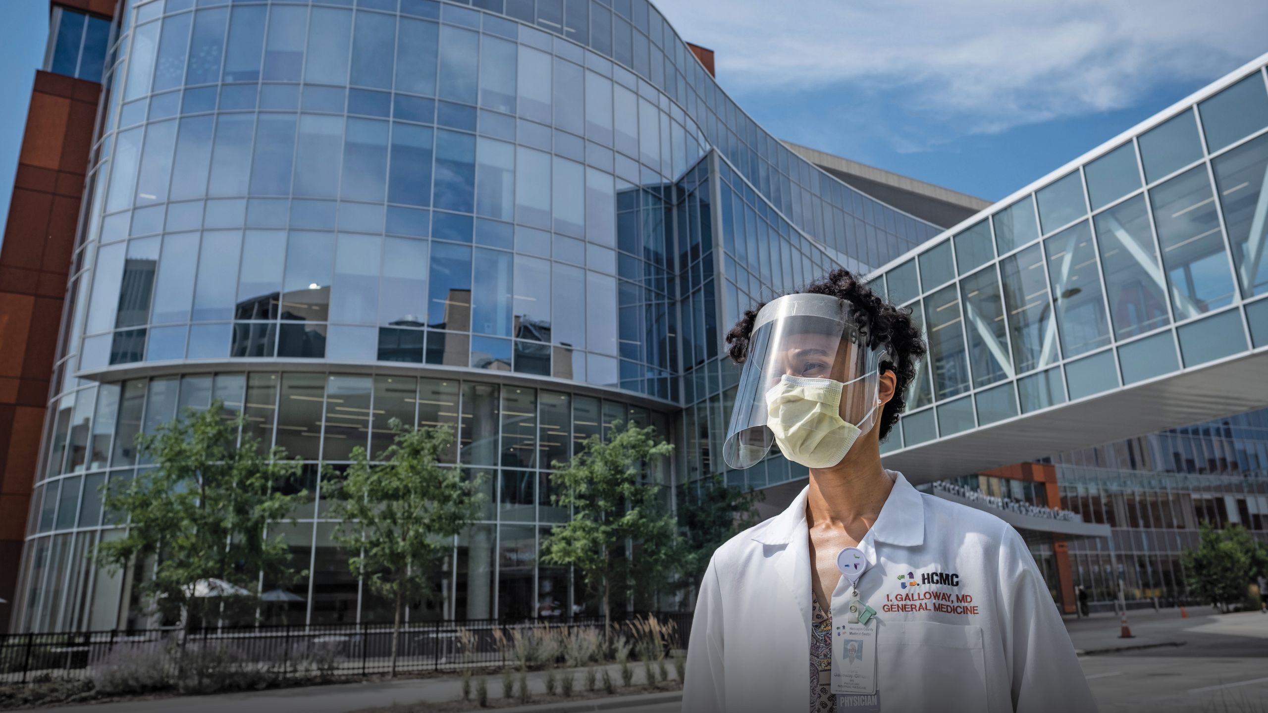 A female doctor stands in front of a hospital in a mask and face shield and her white coat. 