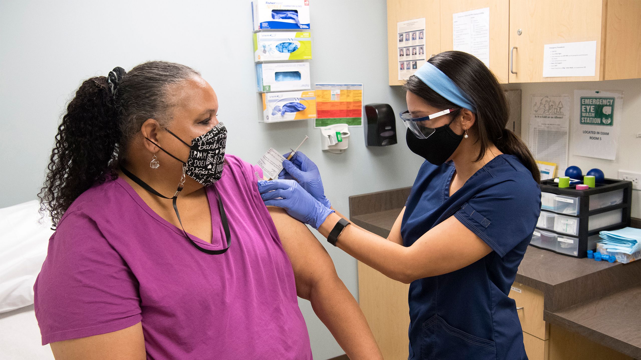 A woman receives an injection in her arm. Both the volunteer and the woman administering the injection are wearing face coverings