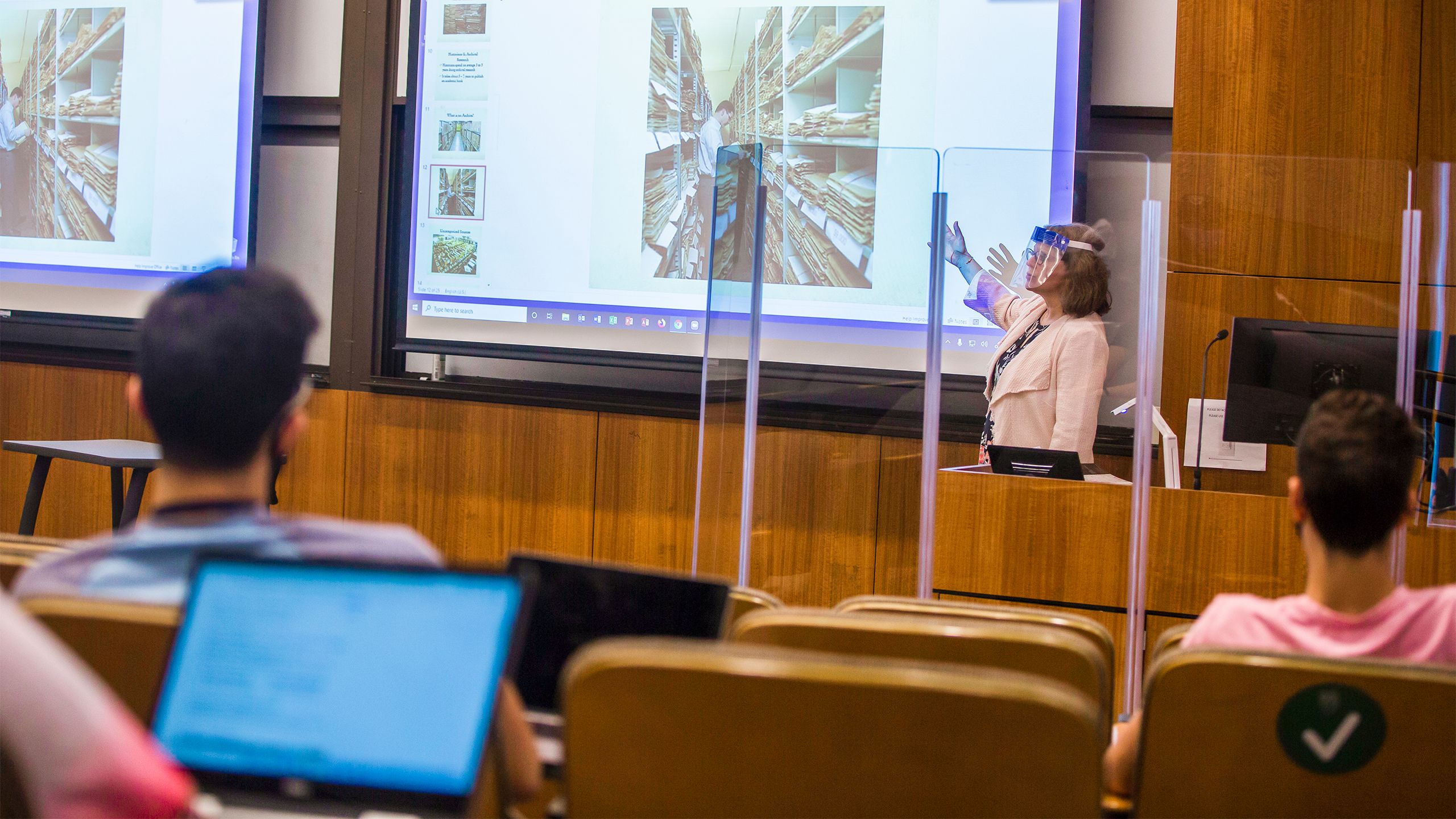 A classroom shows a professor teaching wearing a face shield and standing behind a clear plastic barrier