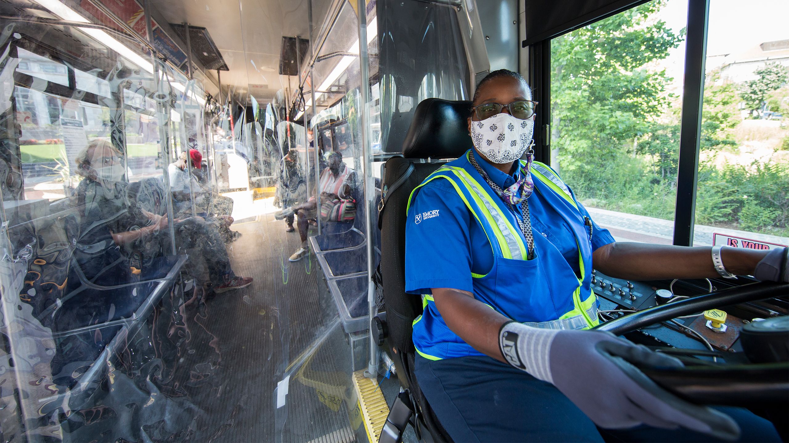 An Emory bus driver sits behind the wheel wearing gloves and a face covering. Passengers are also sitting far apart and wearing face coverings