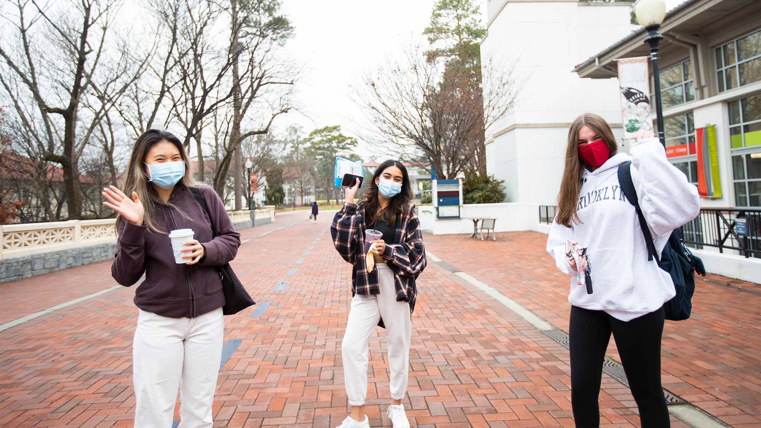 Three students wearing face coverings and sweatshirts wave from the bridge in front of Cox Hall