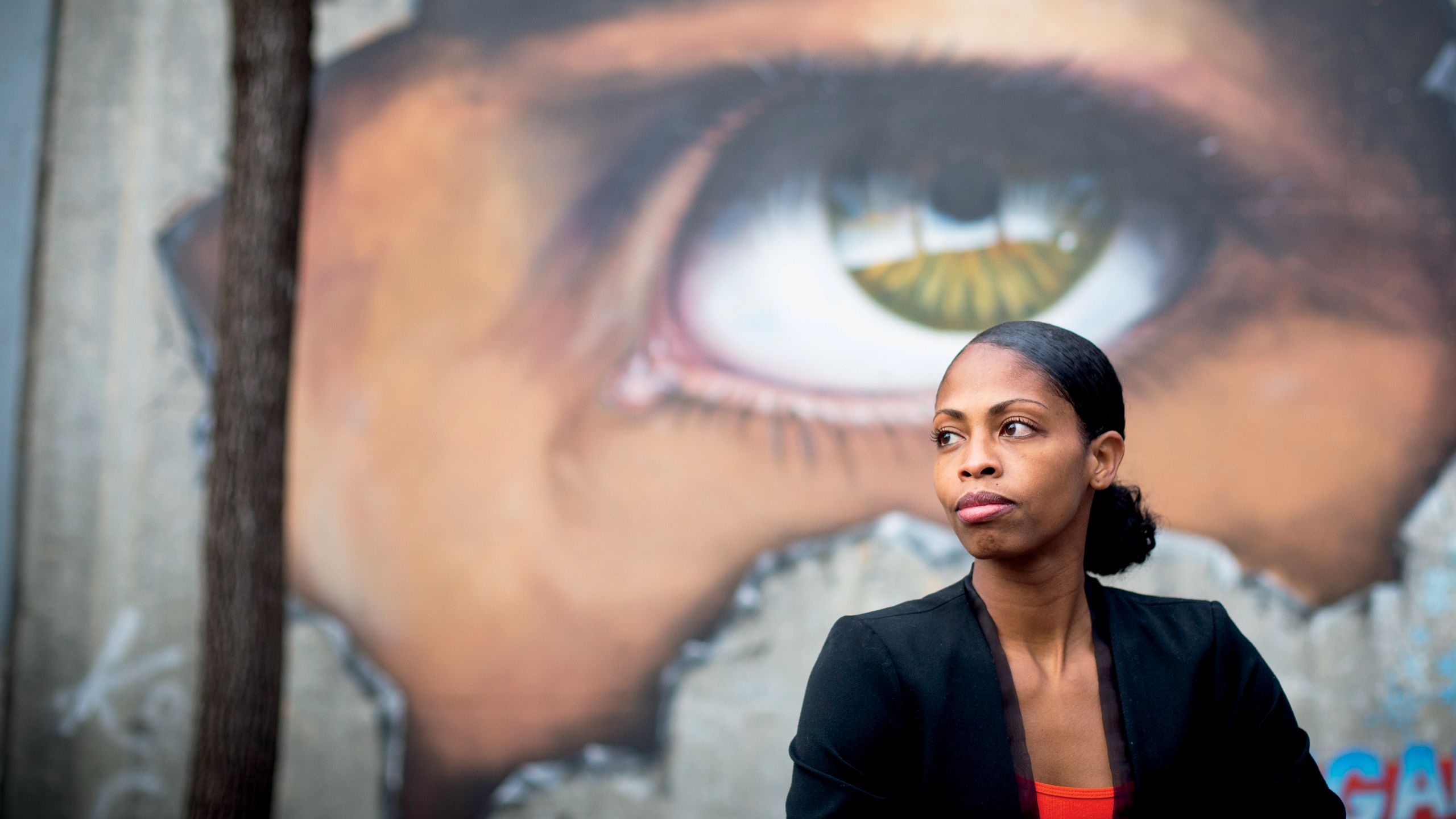 a photo of Lauren McCullough posing in front of a mural by Julio Ceballos. The mural depicts a large eyeball peering through a cracked wall.