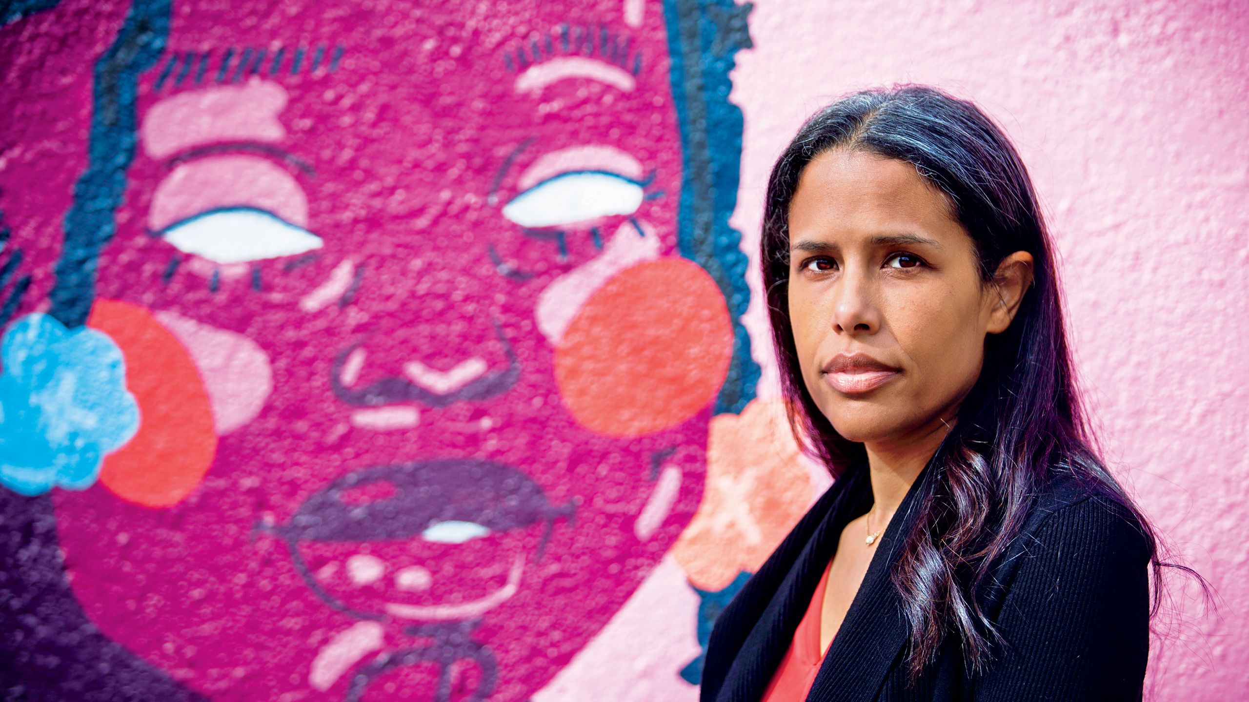 a photo of Briana Woods-Jaeger standing in front of a mural by Austin "Blue" Richardson. The mural depicts a young, black girl who passed away due to gun violence. 