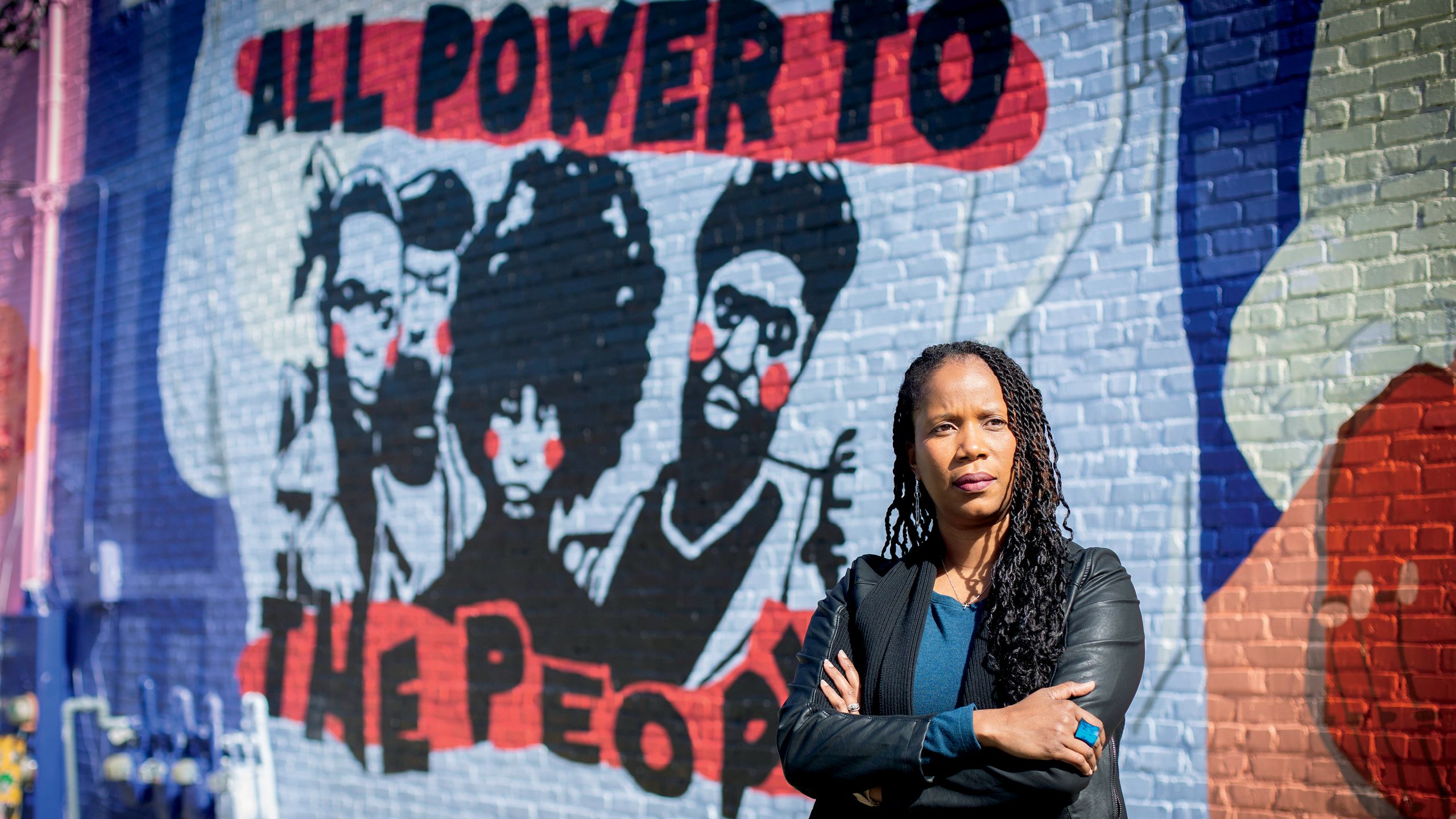a photo of dr. dana williamson standing in front of a mural. The mural has a white shirt with a group of people with the text that reads all power to the people. There is also a mural of a young black child right next to that. The mural is by SofaHood.