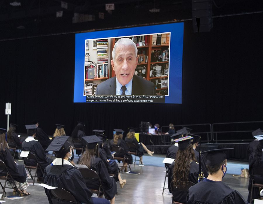 Students in caps and gowns watch a large screen with Dr. Anthony Fauci