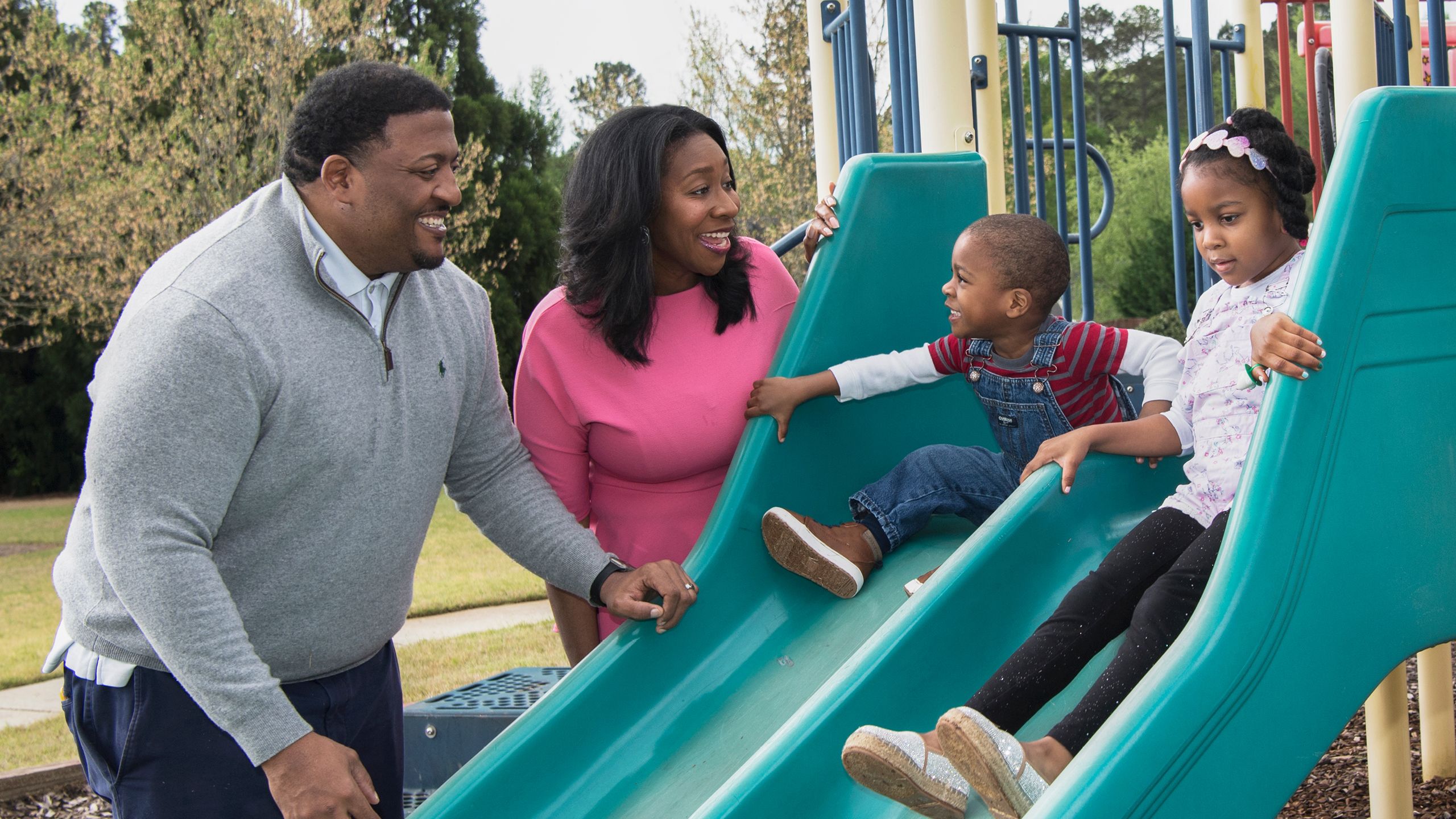 A photograph of a family in a park, with a young boy on a slide, a slightly older girl beside him on another slide, and their mom and dad smiling and standing beside them watching them. 