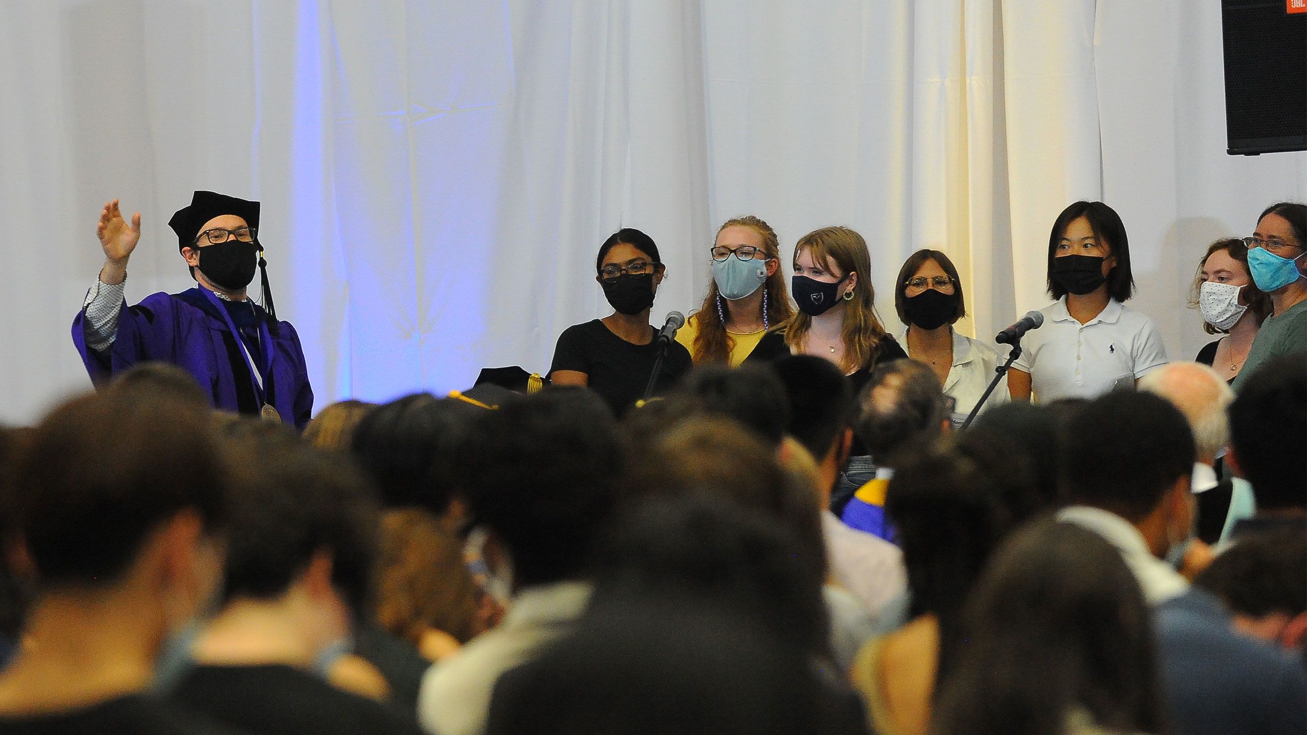 Students stand at a microphone in masks to sing the alma mater