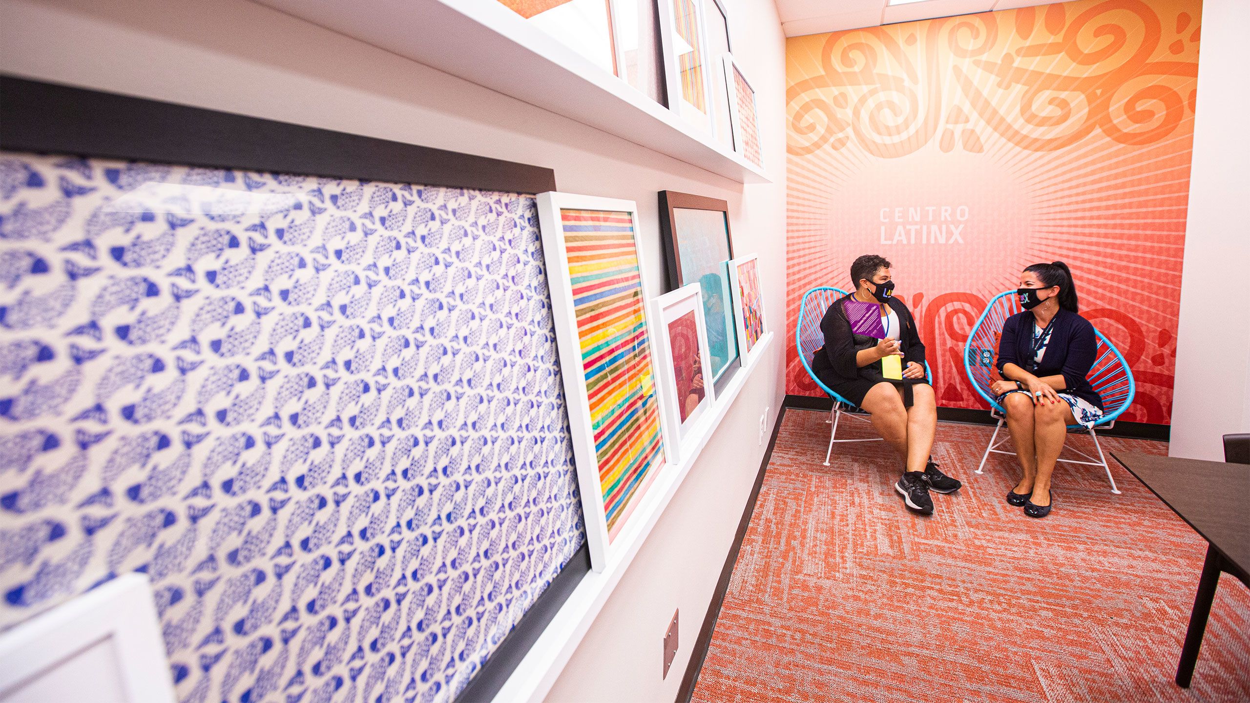 Two people sit in chairs in front of a brightly colored mural in Centro Latinx