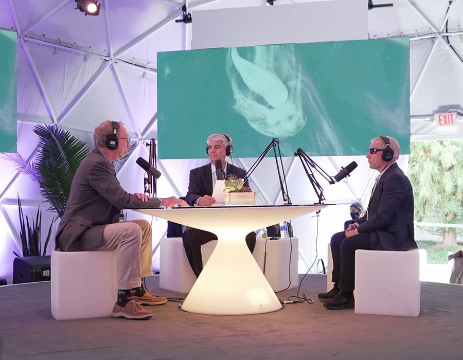 Emory President Gregory L. Fenves (center) hosts a discussion about drug discovery and development with professors George Painter (left) and Dennis Liotta, sitting at a lighted table with microphones and headphones.