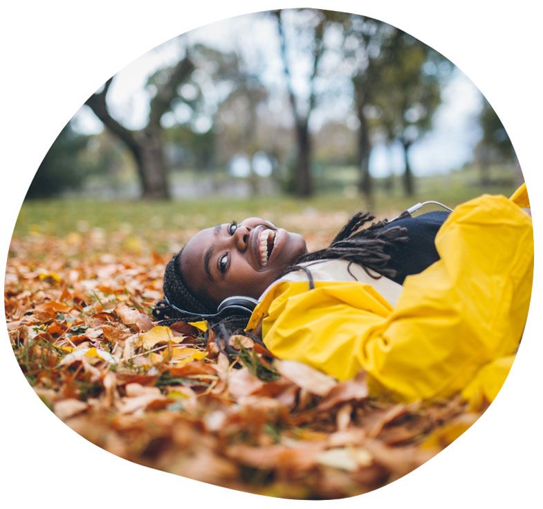 A student listening to headphones lies on the ground smiling and surrounded by fall leaves.