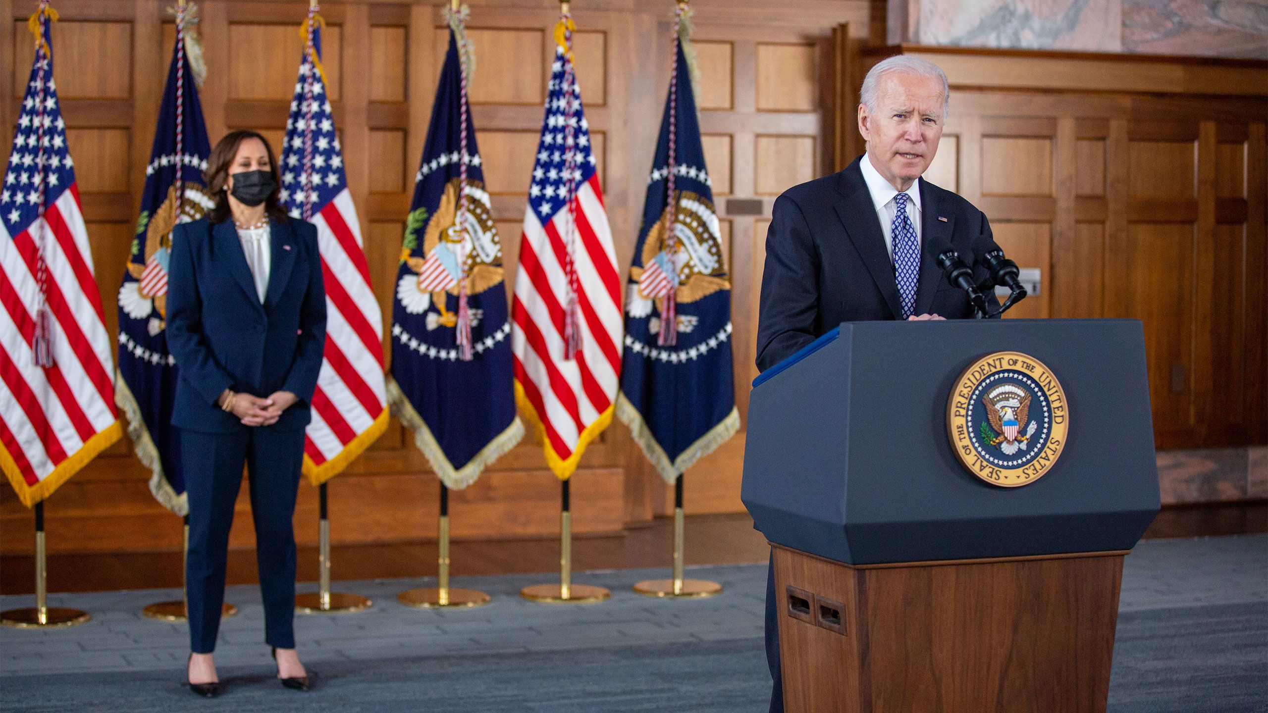 President Joe Biden speaks at a podium in Emory's Convocation Hall with a row of flags behind him. Vice President Kamala Harris stands in front of the flags.
